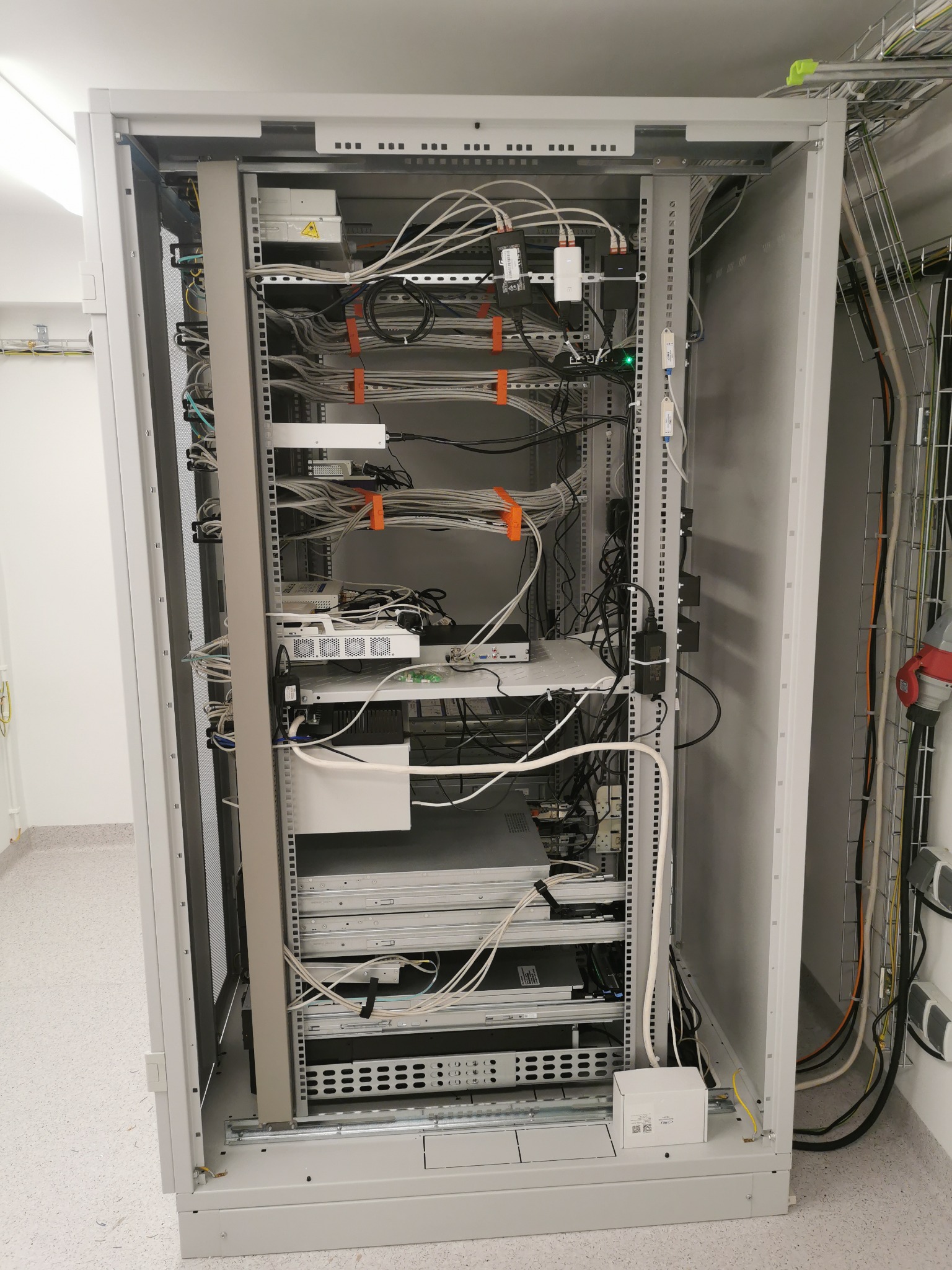 Otwarta szafa serwerowa z zainstalowanymi urządzeniami sieciowymi, widoczne kable uporządkowane pomarańczowymi opaskami, zasilacze UPS i listwy zasilające, okablowanie prowadzone w korytkach...