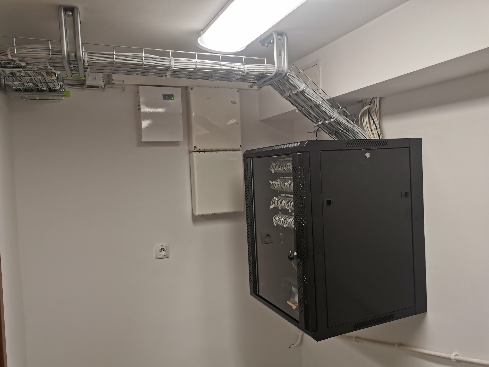 Kable w korytach siatkowych pod sufitem, szafa rack z widocznym okablowaniem wewnątrz, białe ściany i gniazdko elektryczne