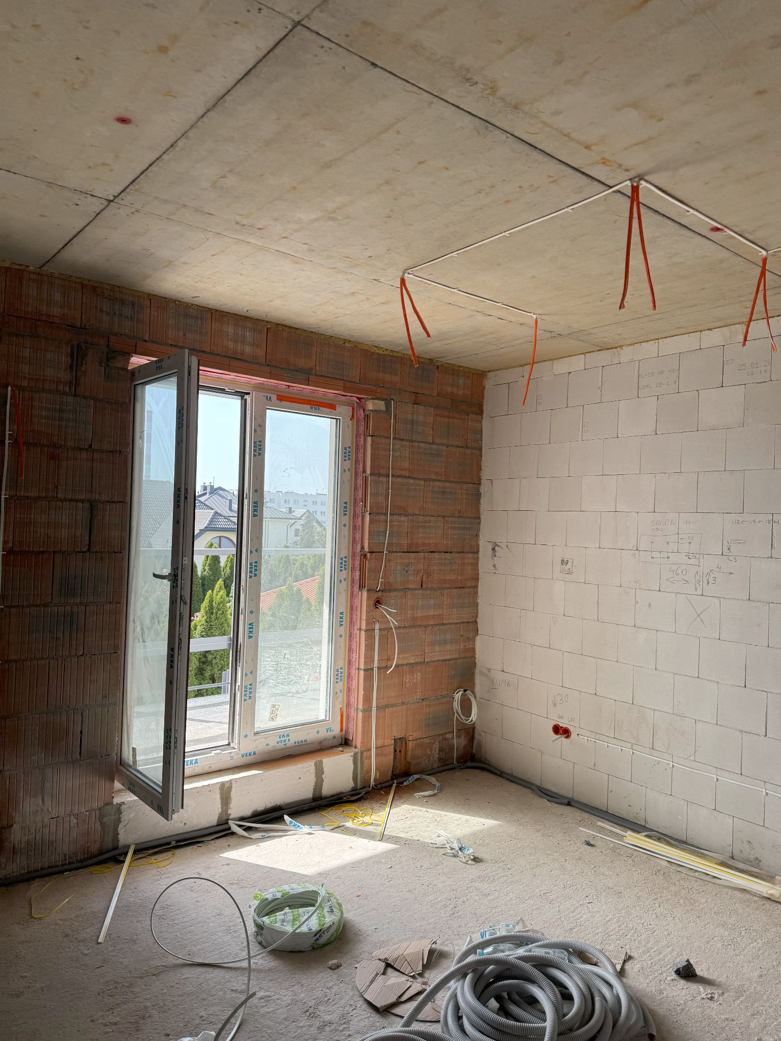 Wnętrze surowego domu w trakcie prac instalacyjnych, widoczne kable elektryczne pod sufitem mocowane pomarańczowymi paskami, instalacja okienna z widokiem na zabudowę mieszkalną, oraz zwój szarego...