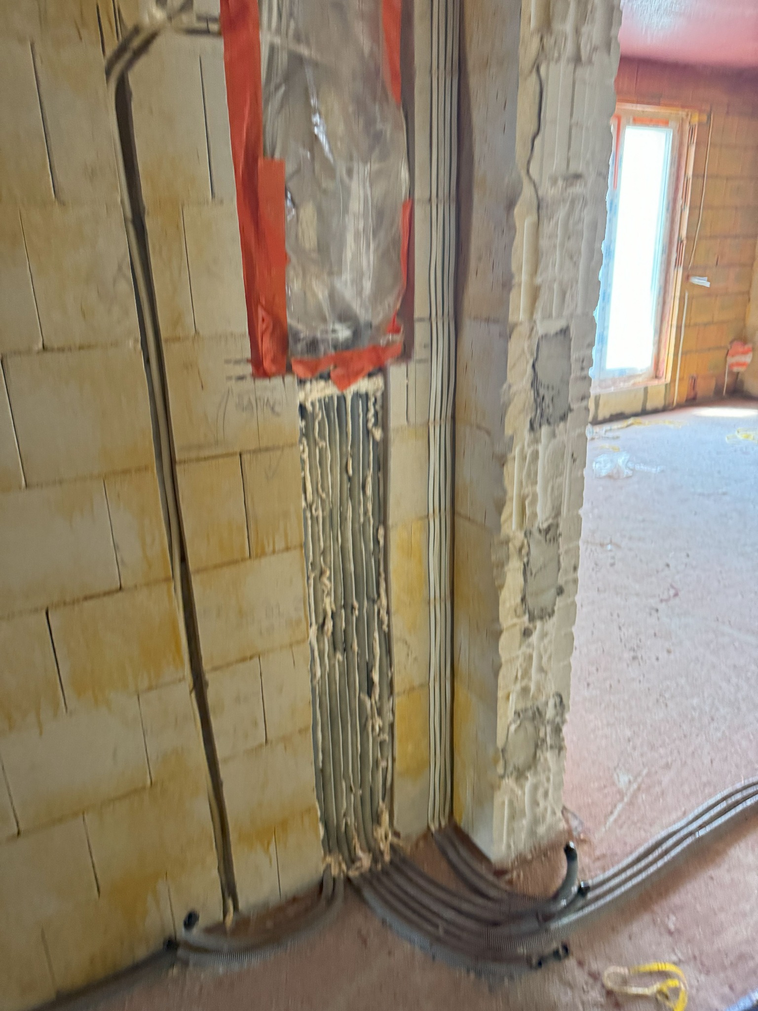 Wykute bruzdy w ścianie z betonu komórkowego, wypełnione peszlami z kablami zabezpieczonymi pianką montażową i folią, widoczne kable prowadzone po podłodze.