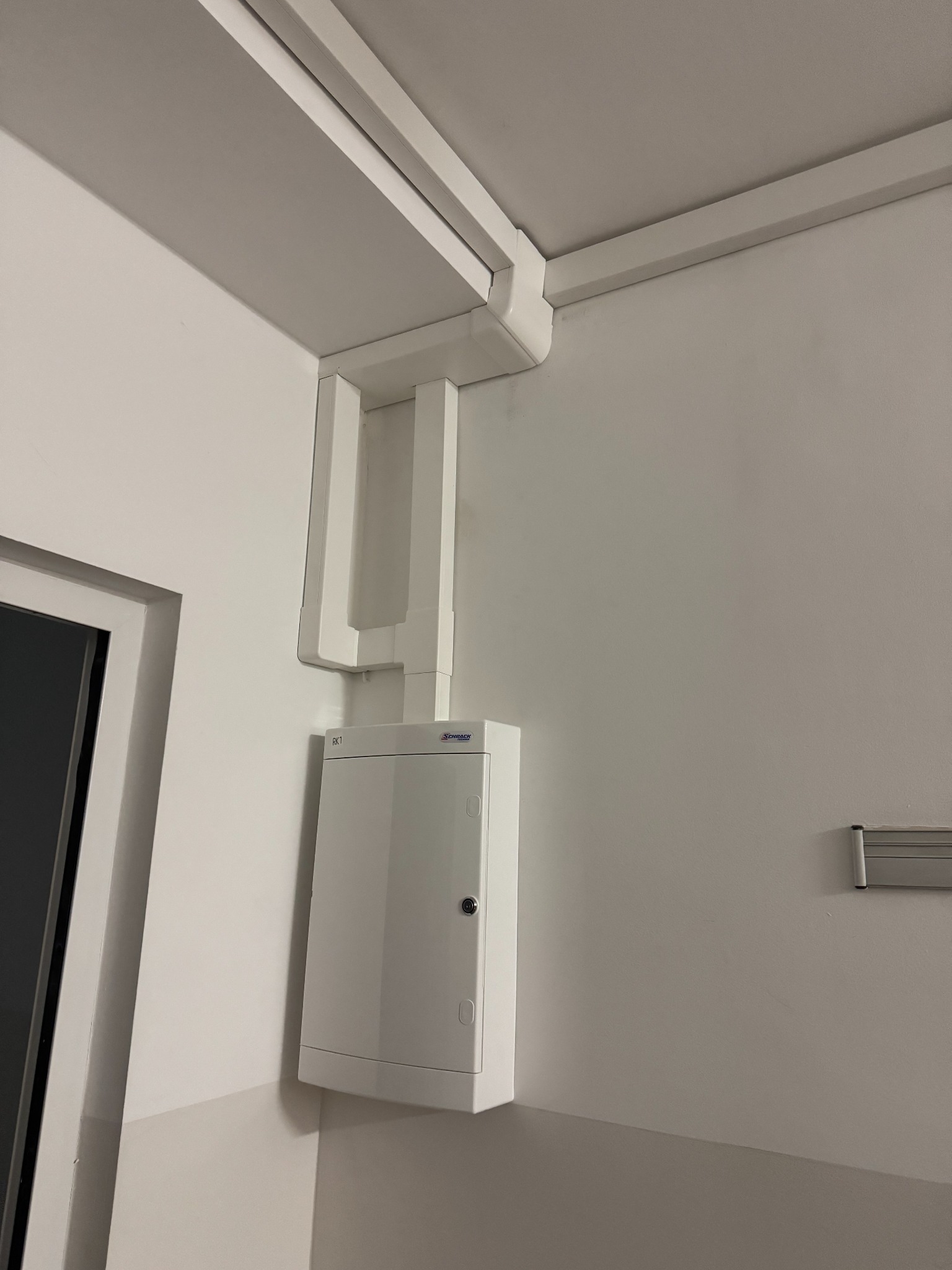 Biała skrzynka elektryczna zamontowana na ścianie w rogu pomieszczenia, z widocznymi białymi rurami prowadzącymi do sufitu, obok framugi okna.