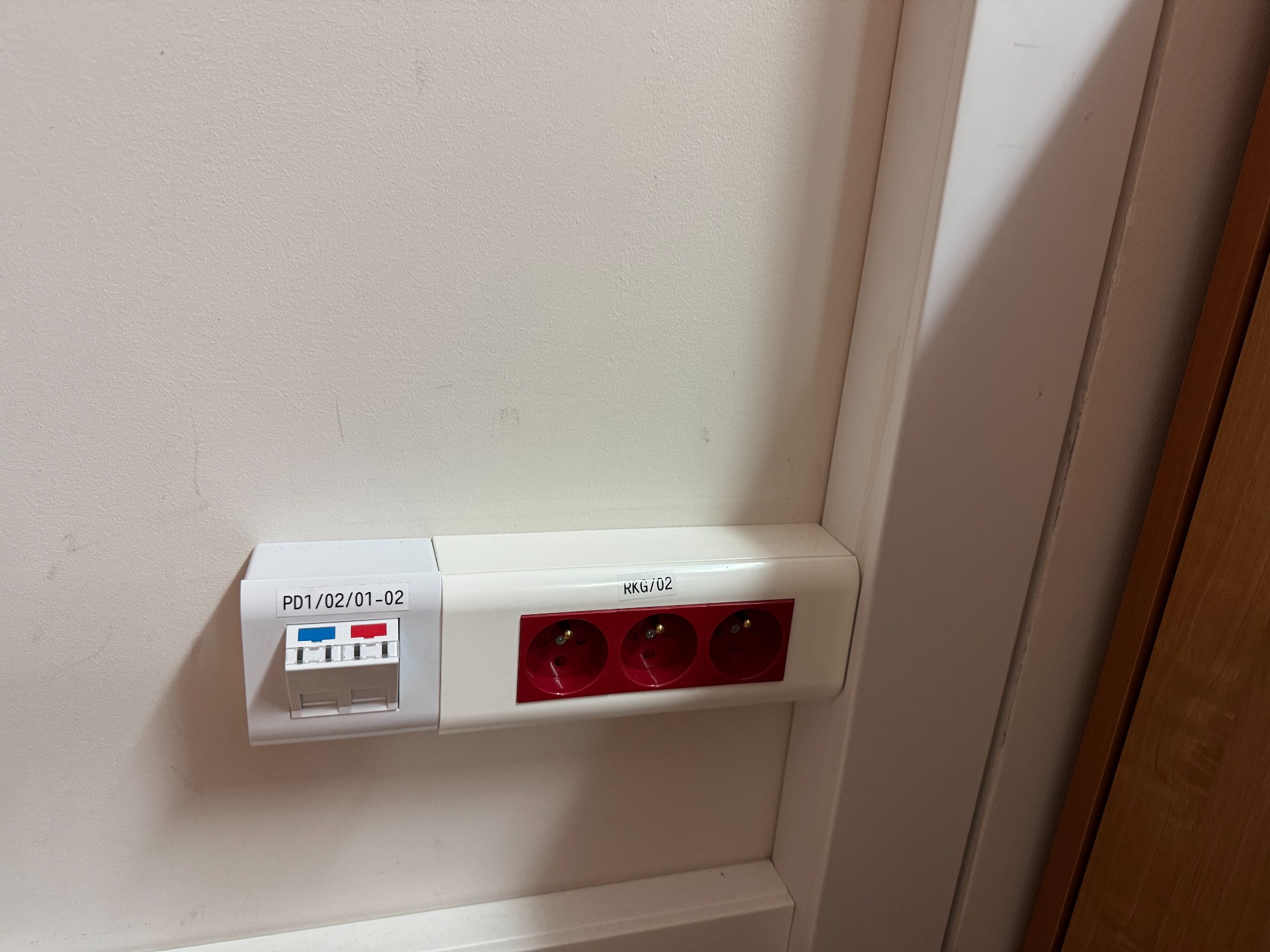 Biała listwa przypodłogowa z trzema czerwonymi gniazdami elektrycznymi i zabezpieczeniem nadprądowym z oznaczeniami PD1/02/01-02 i RKG/02, zamontowana na jasnej ścianie w pobliżu drewnianej framugi.