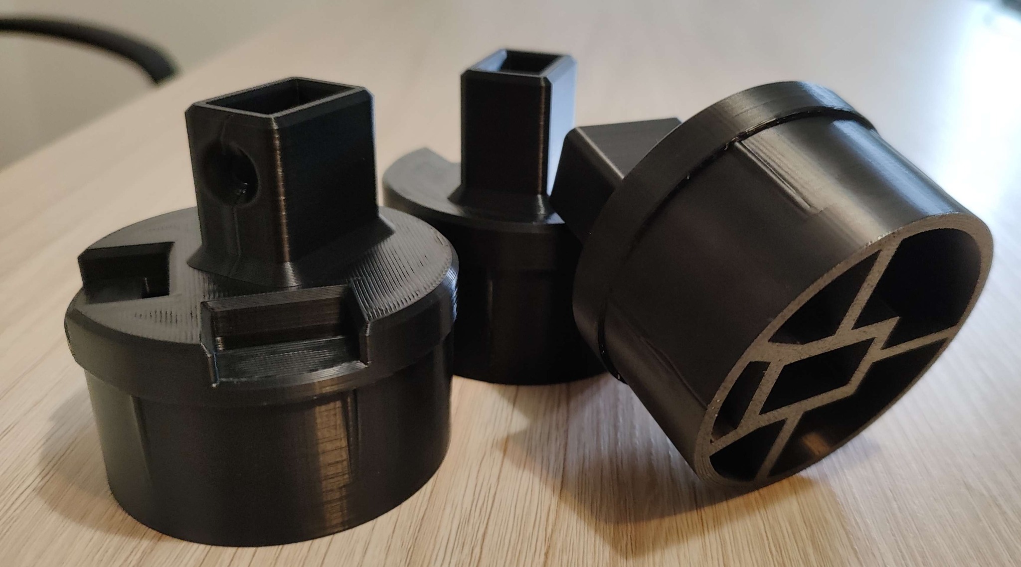 Trzy czarne, precyzyjnie wykonane elementy z tworzywa sztucznego z widoczną strukturą warstwową, leżące na jasnym, drewnianym blacie. Różne kształty i detale.