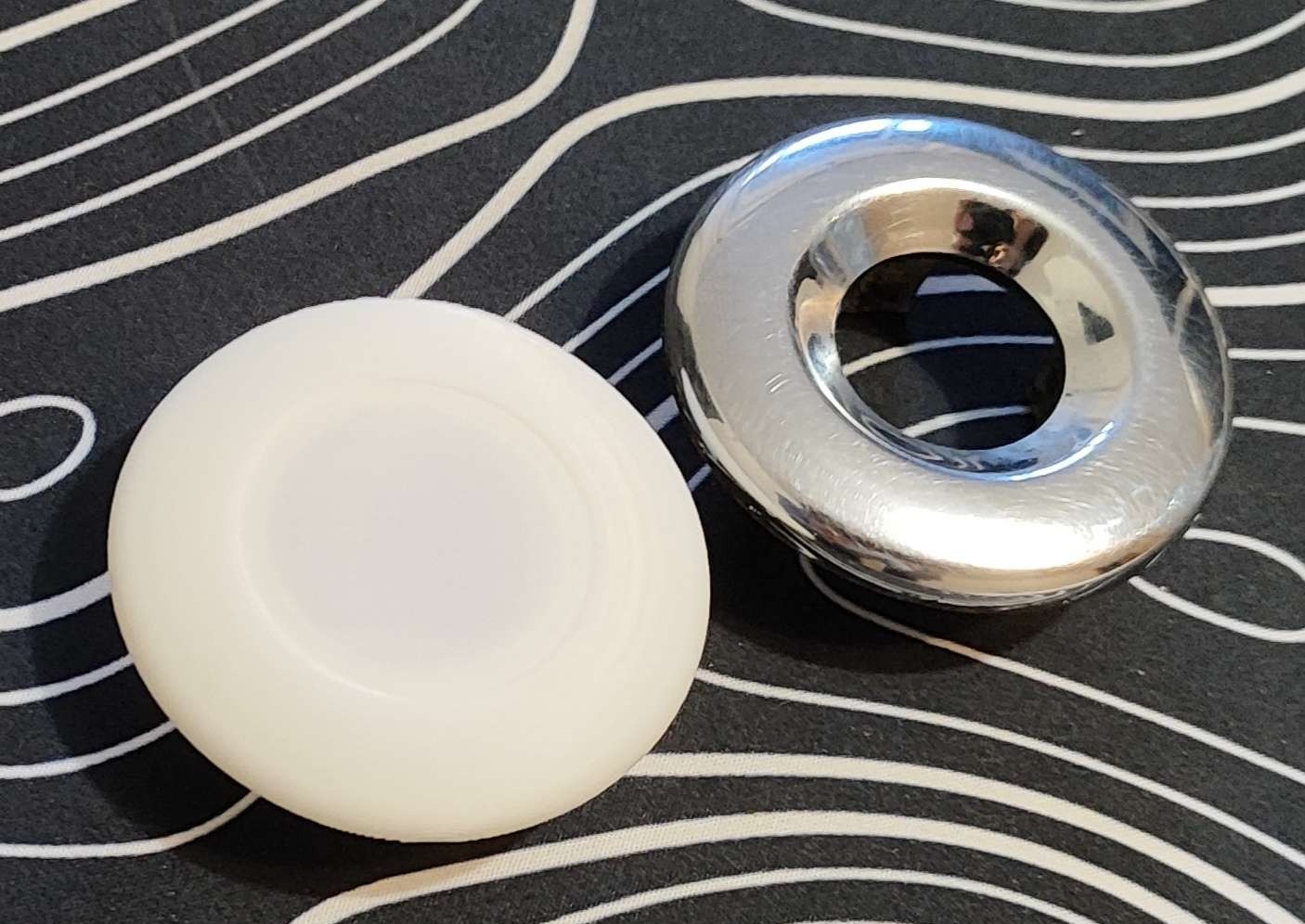 Biały i srebrny element z otworem, wykonane metodą addytywną, leżą na czarno-białym tle w paski. Elementy o gładkiej powierzchni, widoczne delikatne warstwy.