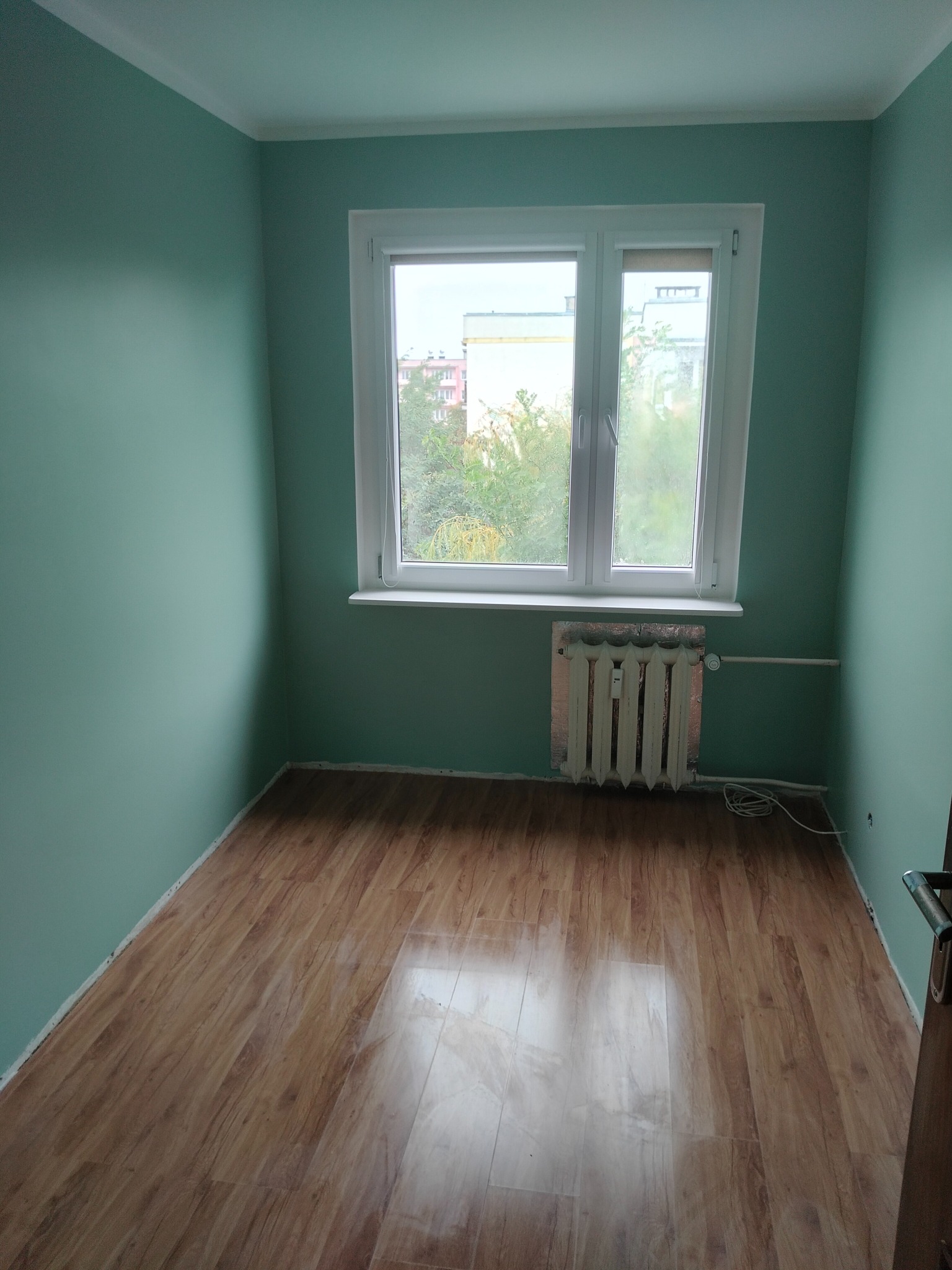 Pomalowane na miętowo wnętrze pokoju z panelami podłogowymi, białym oknem i grzejnikiem z folią termoizolacyjną. Widok na zieleń za oknem.