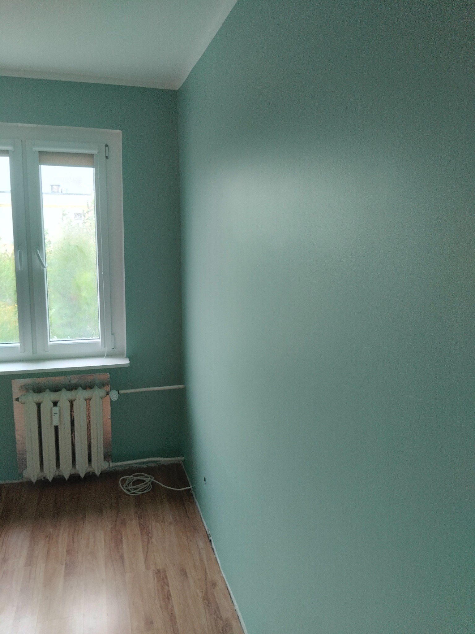 Ściana pomalowana na jednolity, pastelowy odcień zieleni w pokoju z drewnianą podłogą, białym oknem i grzejnikiem. Widoczne niedoskonałości malowania przy grzejniku.