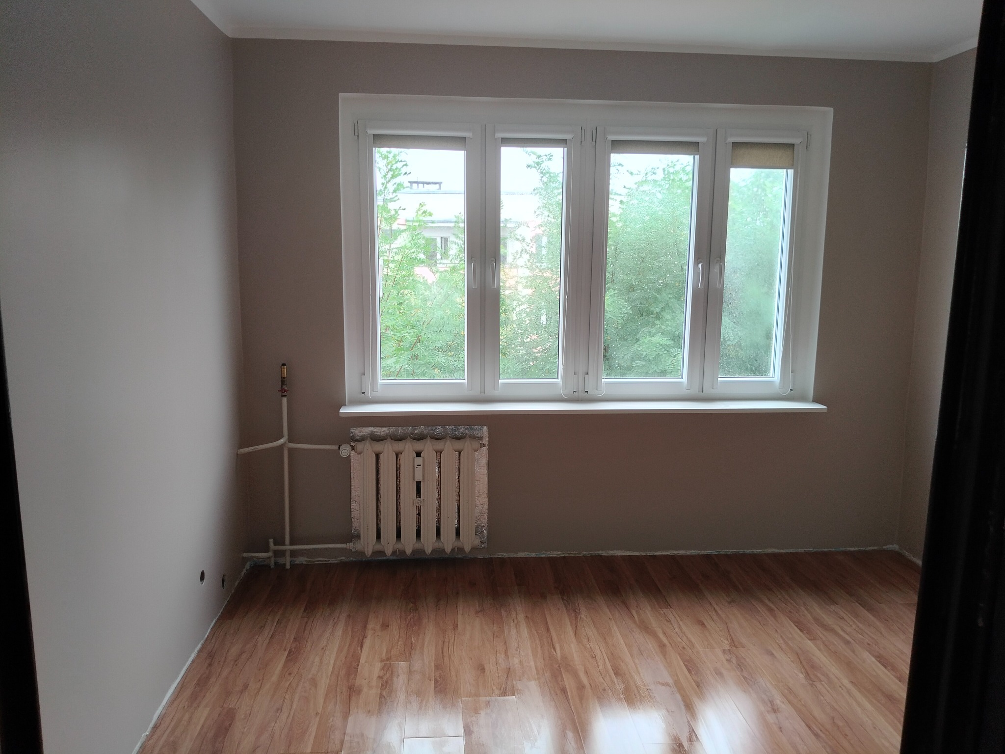 Pusty pokój po remoncie: świeżo pomalowane ściany w odcieniach beżu, panele podłogowe imitujące drewno, białe okno z widokiem na zieleń, grzejnik z folią termoizolacyjną.