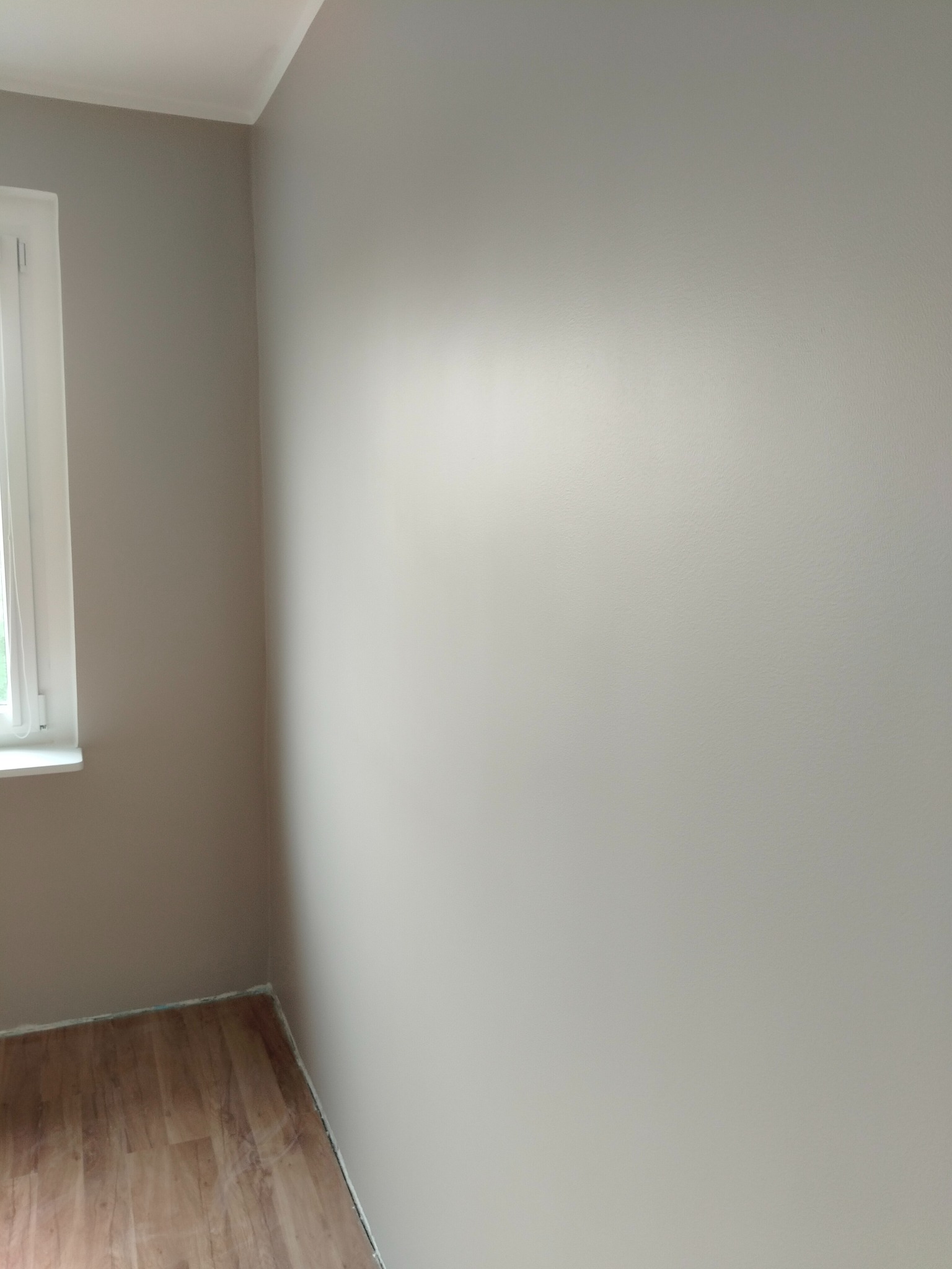 Jasnoszary pokój po malowaniu z drewnianą podłogą i białym oknem. Świeżo pomalowane ściany, widoczny fragment podłogi i okno z parapetem.