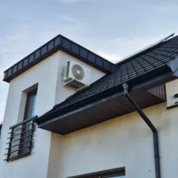 Biała jednostka zewnętrzna klimatyzacji zamontowana na elewacji nowoczesnego domu z ciemną dachówką, widok z dołu na tle jasnego nieba.