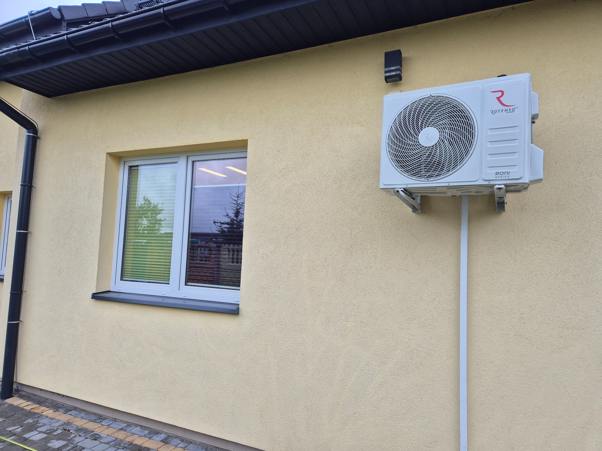 Biała klimatyzacja Rotenso Roni Series zamontowana na elewacji domu o beżowej strukturze, obok okna z odbijającym się drzewem. Widoczny fragment dachu i rynna.