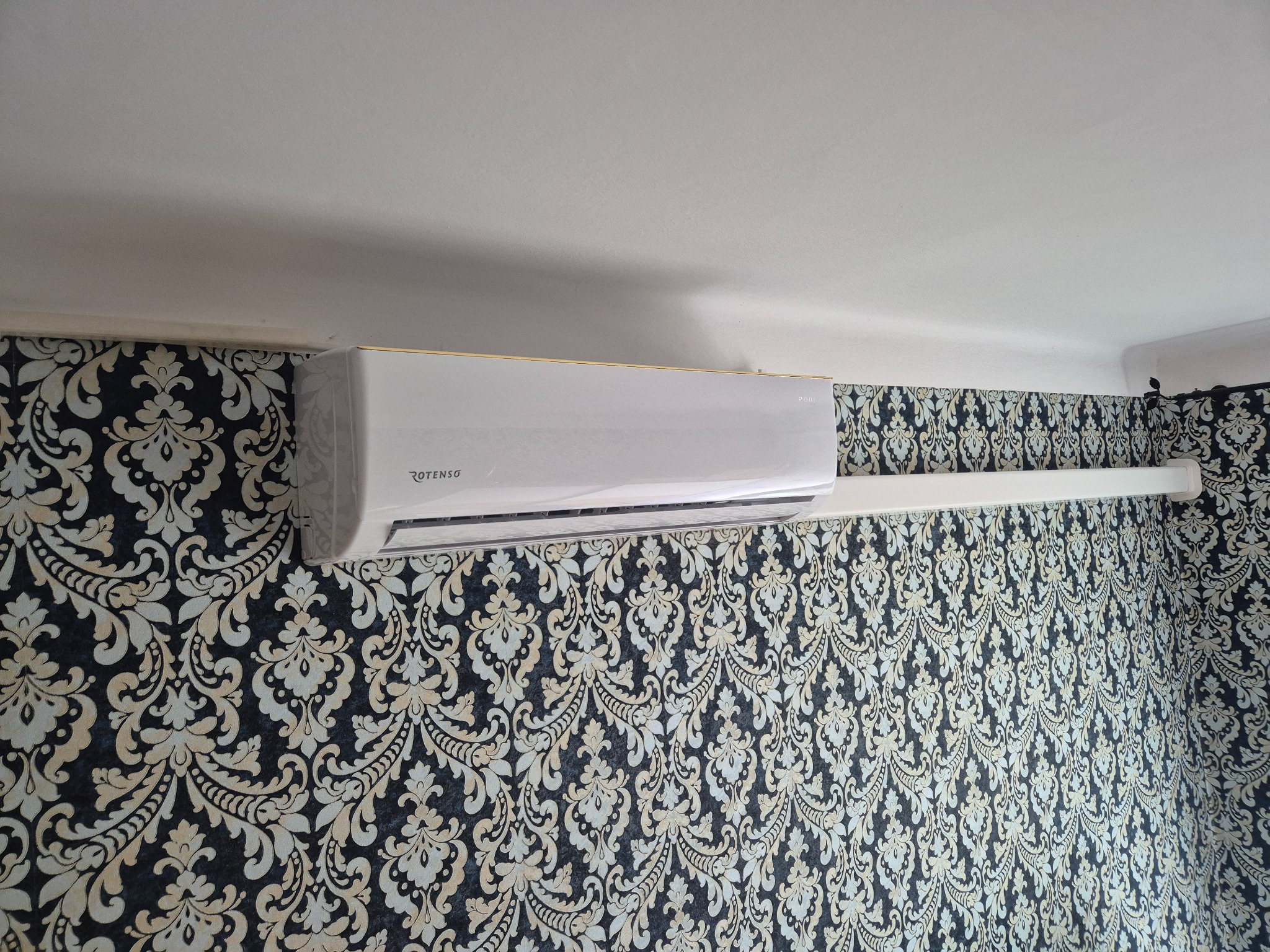 Biały klimatyzator Rotenso na ścianie z wzorzystą tapetą. Widok z dołu,  wystrój wnętrza.