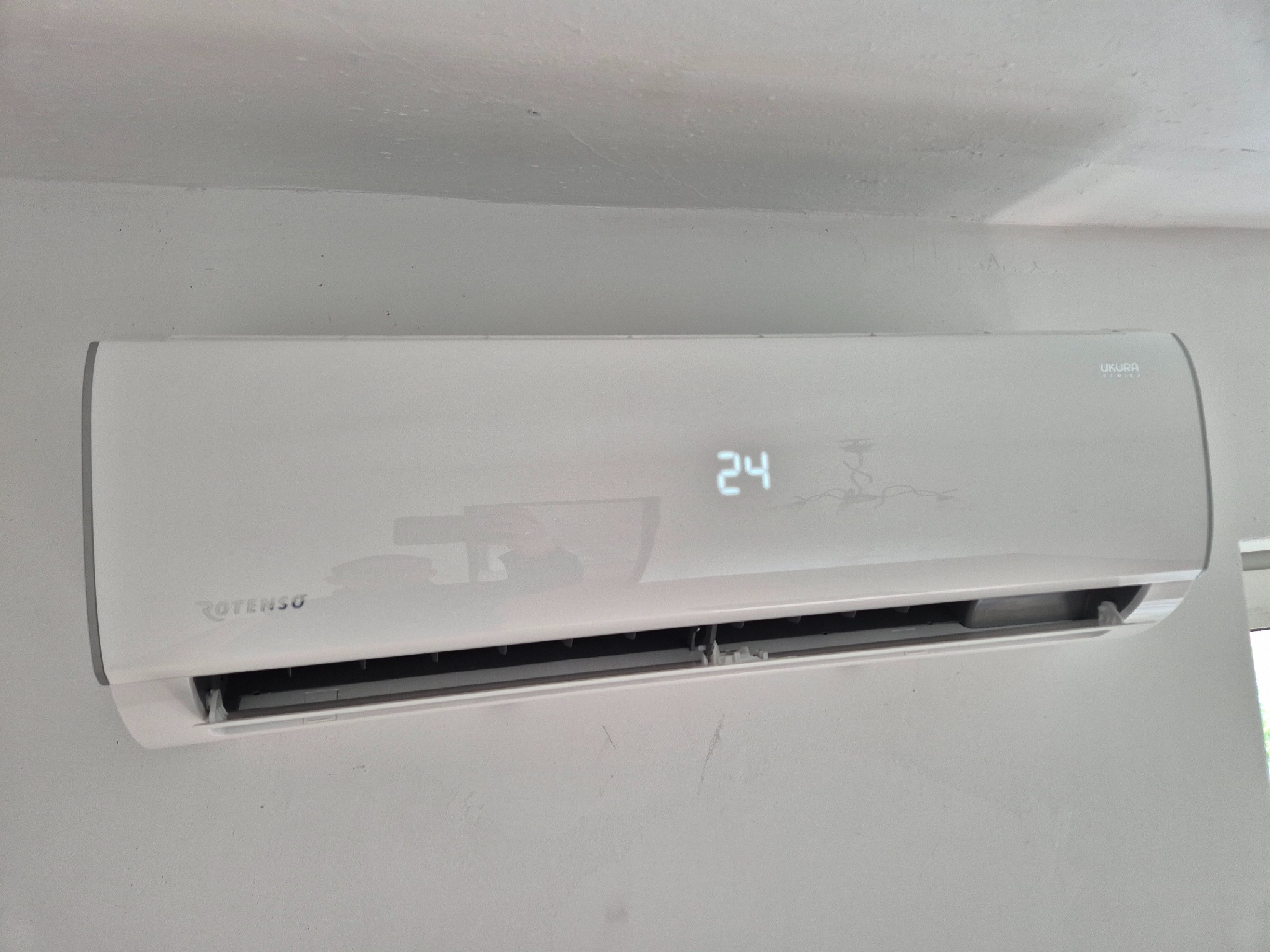 Biała klimatyzacja Rotenso Ukura Series zamontowana na ścianie, wyświetlająca temperaturę 24 stopnie. Widoczny fragment okna po prawej stronie.