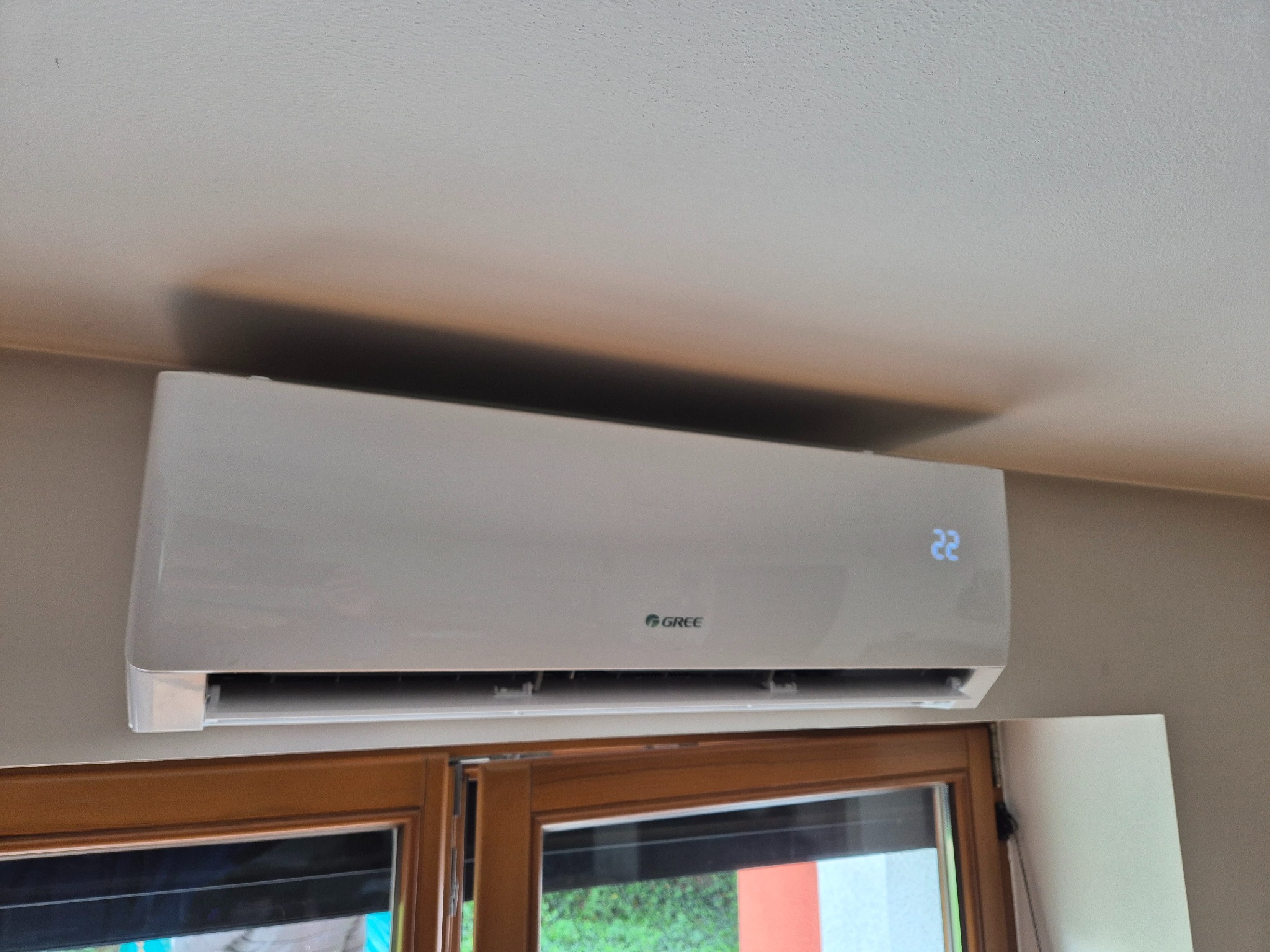 Biały klimatyzator Gree zamontowany nad drewnianym oknem, wyświetlający temperaturę 22 stopnie, w jasnym wnętrzu. Widok z dołu.