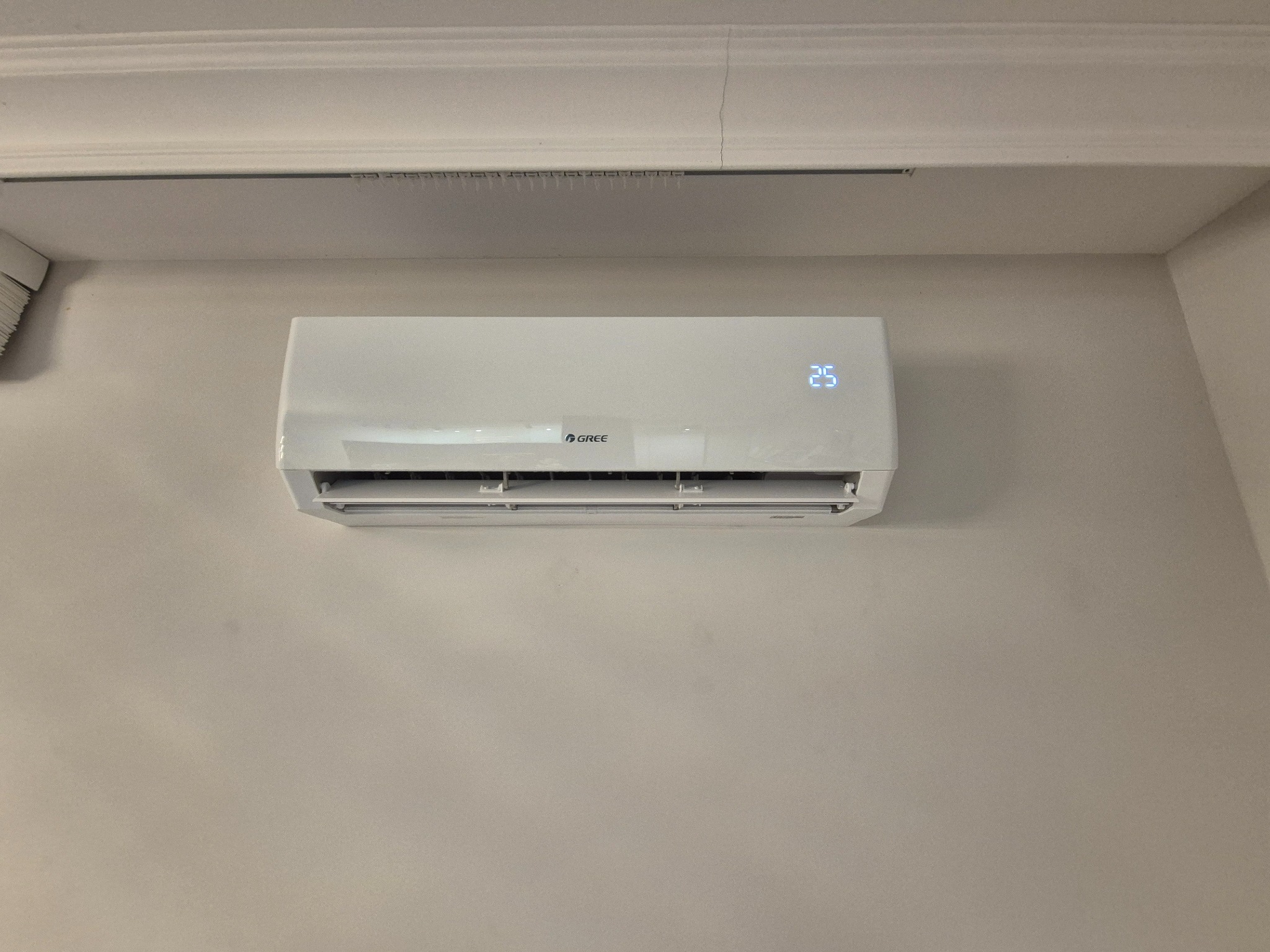 Biała klimatyzacja GREE na ścianie, wyświetlająca temperaturę 25 stopni. Widoczne listwy sufitowe i fragment żaluzji w lewym górnym rogu.