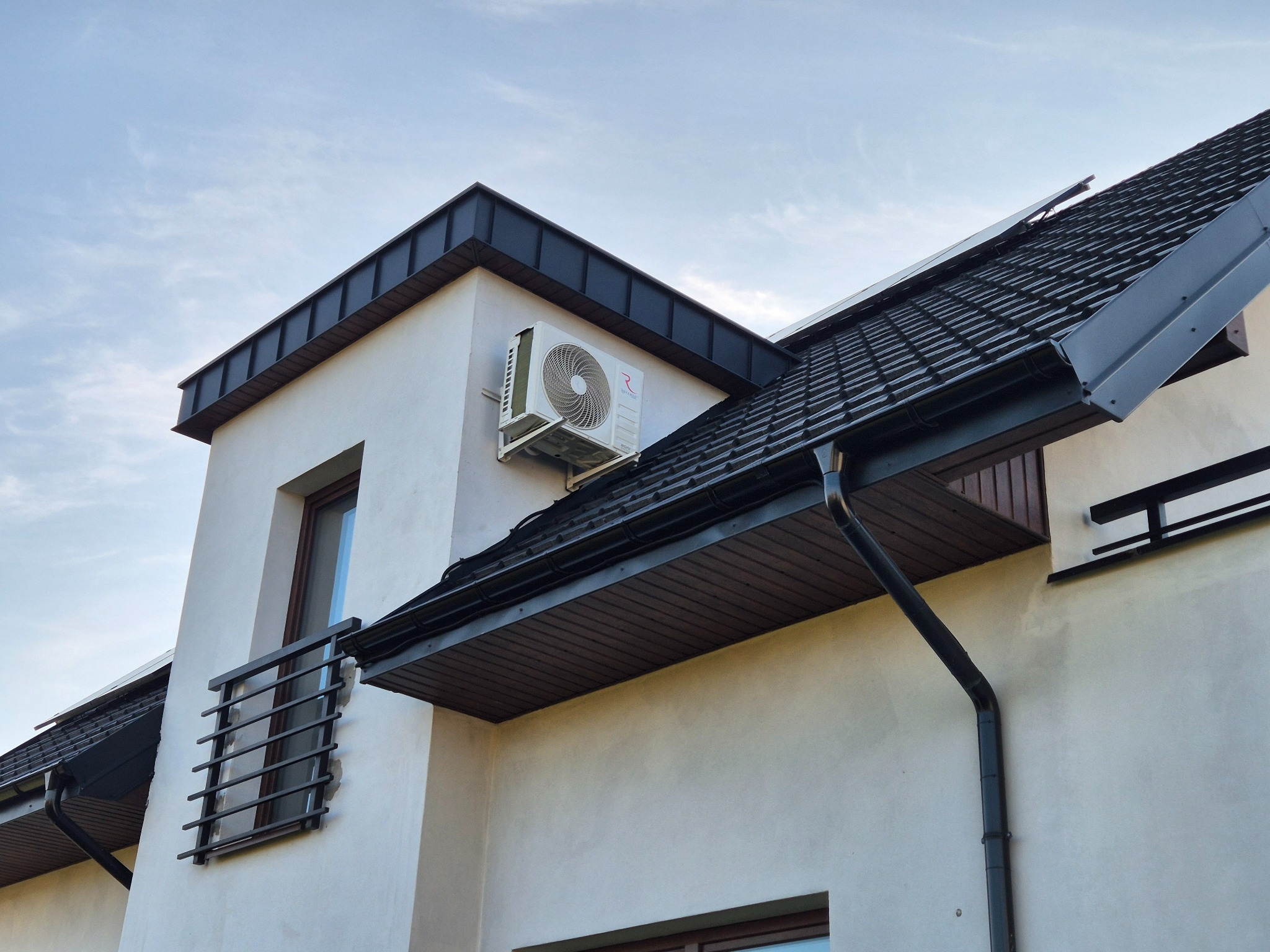 Biała jednostka zewnętrzna klimatyzacji zamontowana na elewacji nowoczesnego domu z ciemną dachówką, widok z dołu na tle jasnego nieba.