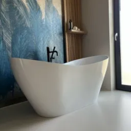 Wolnostojąca biała wanna na jasnej posadzce w łazience z akcentem turkusowych piór na ścianie i drewnianymi lamelami. Czarna bateria wannowa. Nowoczesny design.