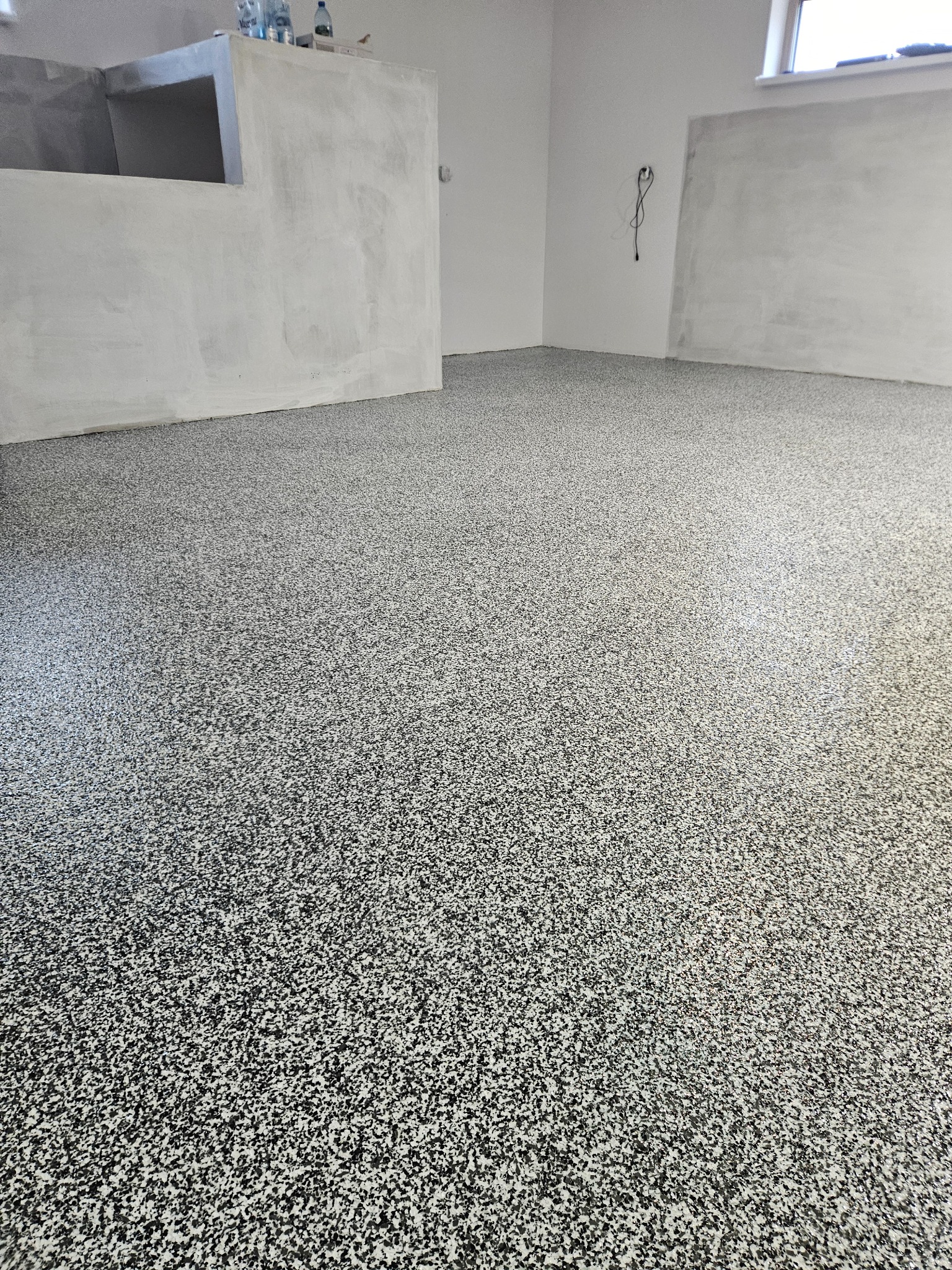 Wykończona podłoga w pomieszczeniu z surowymi ścianami, pokryta szaro-biało-czarnym wzorem przypominającym lastryko, widoczne butelki z wodą na betonowej konstrukcji w tle.