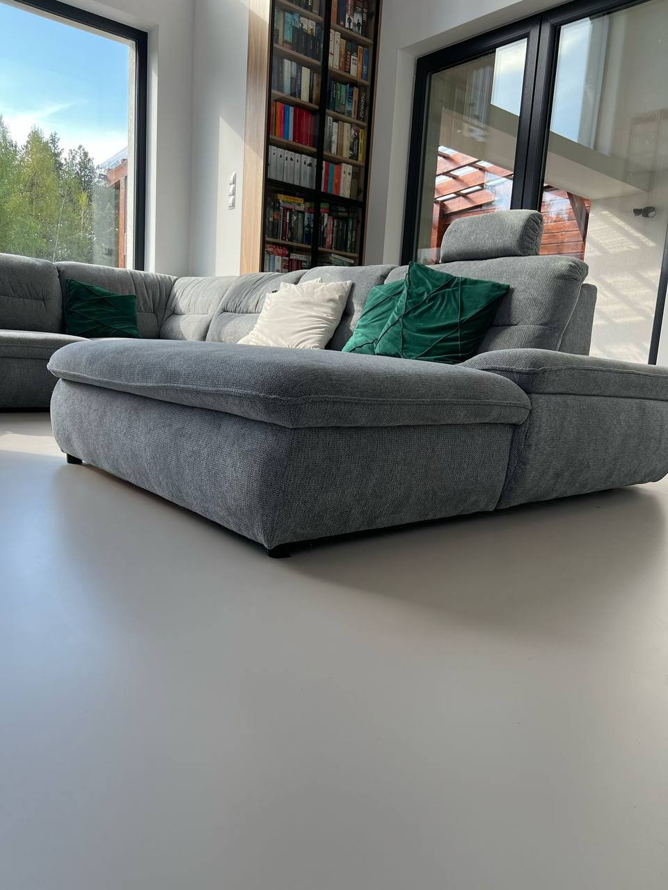 Szara, modułowa sofa z zielonymi poduszkami na gładkiej, jasnoszarej posadzce w nowoczesnym wnętrzu z dużymi oknami i regałem na książki.