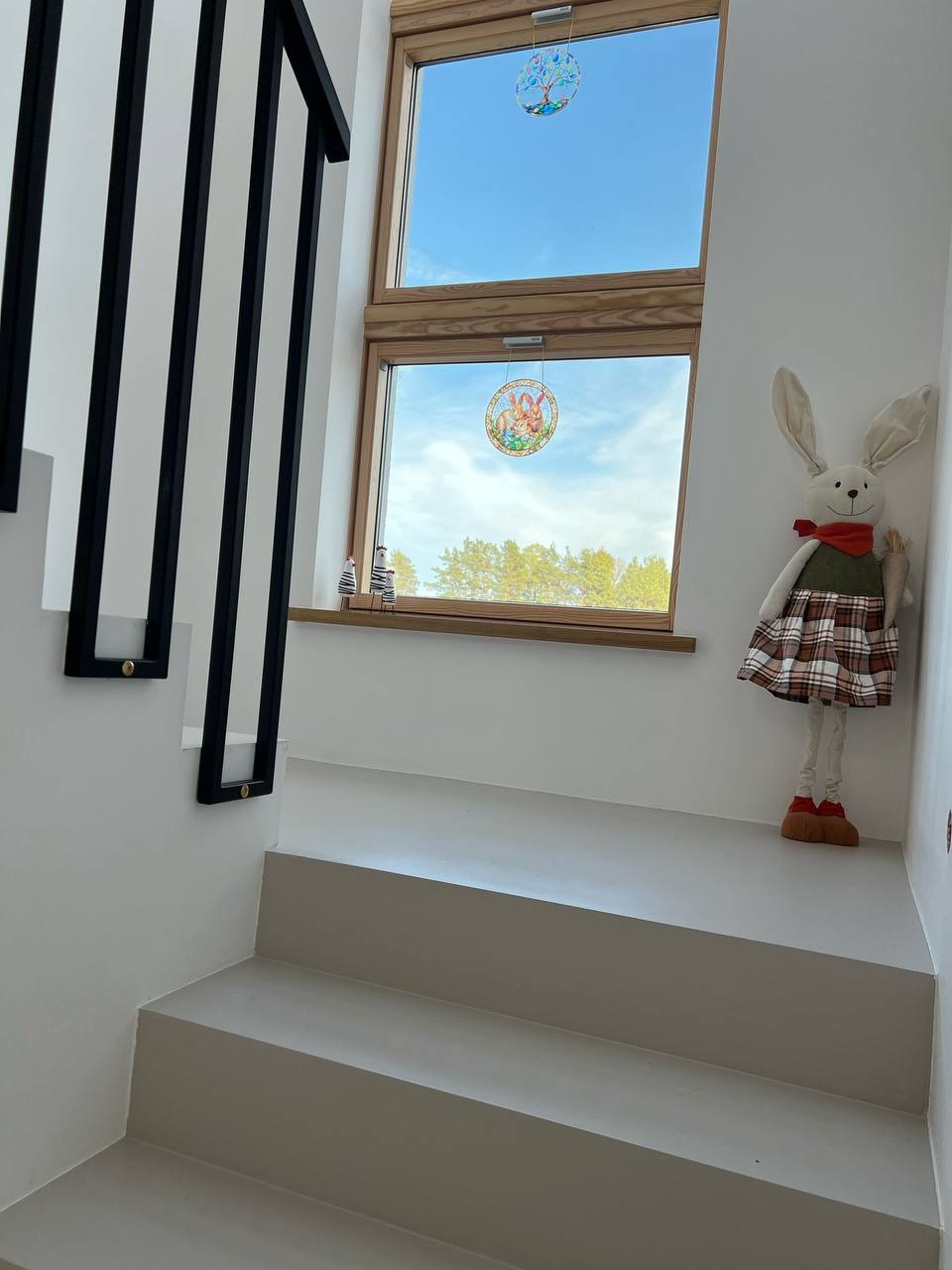 Minimalistyczne schody z jasną, jednolitą posadzką żywiczną, czarne balustrady i okno z dekoracjami, pluszowy królik jako akcent dekoracyjny.