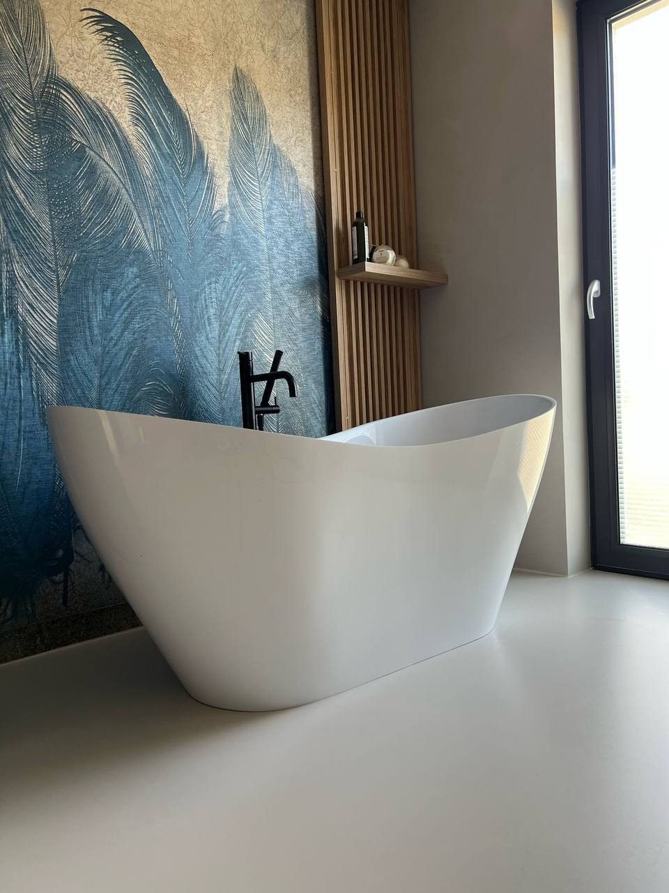 Wolnostojąca biała wanna na jasnej posadzce w łazience z akcentem turkusowych piór na ścianie i drewnianymi lamelami. Czarna bateria wannowa. Nowoczesny design.
