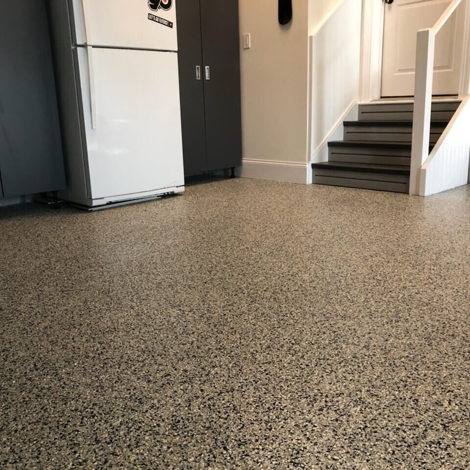 Beżowa posadzka z drobnymi plamkami w garażu, z lodówką i szafkami w tle, schody prowadzące do drzwi. Jasne wnętrze z nowoczesnym wykończeniem.