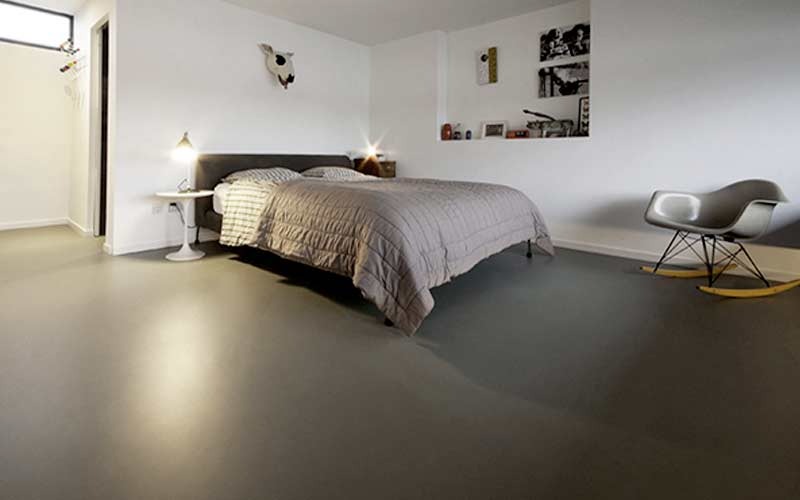 Nowoczesna sypialnia z gładką, szarą podłogą żywiczną. Minimalistyczny design, białe ściany, designerskie krzesło na biegunach, łóżko z szarą pościelą i oświetlenie punktowe.