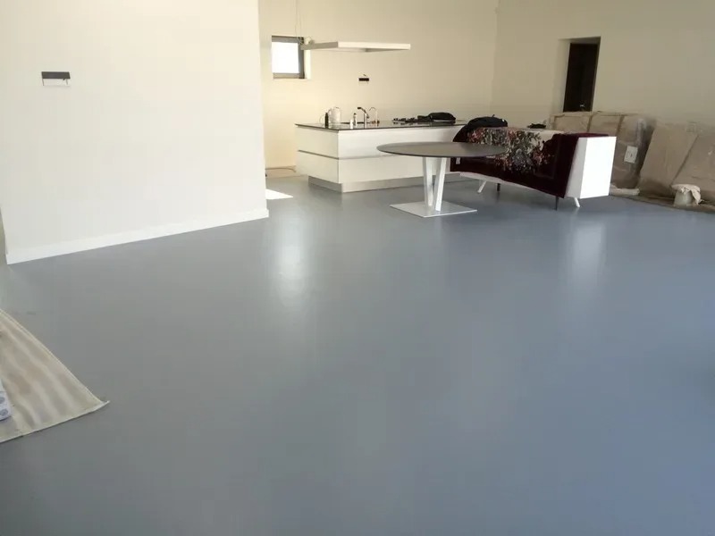 Gładka, szara posadzka w jasnym wnętrzu z otwartą kuchnią. Nowoczesny stół i częściowo zakryte meble w tle. Minimalistyczny design, duże okno zapewnia naturalne światło.