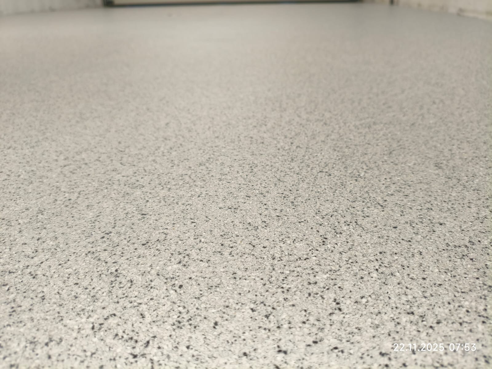 Jasnoszara, gładka posadzka z delikatnym, ciemnym wzorem, sfotografowana pod kątem, ukazująca jednolitą strukturę i brak widocznych łączeń. Minimalistyczny design.