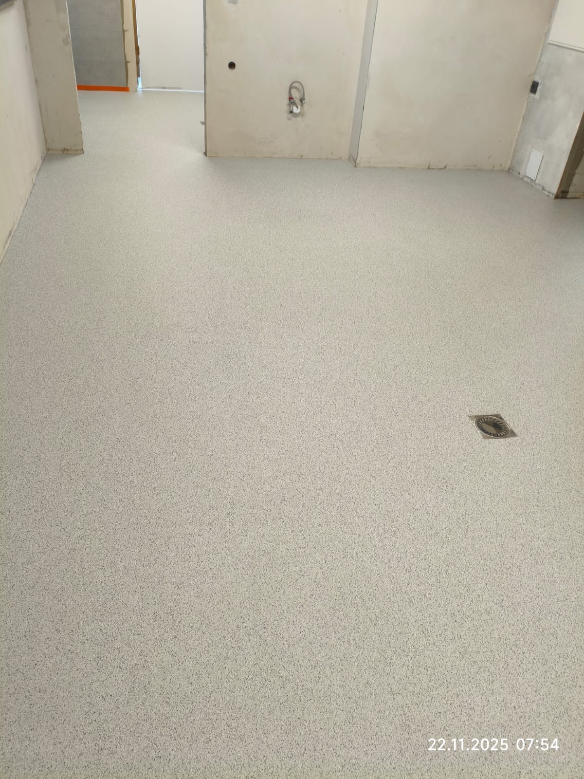 Jasnoszara, drobnoziarnista posadzka w surowym wnętrzu. Widoczny odpływ podłogowy. Ściany w stanie deweloperskim, z widocznymi instalacjami. Minimalistyczny design.