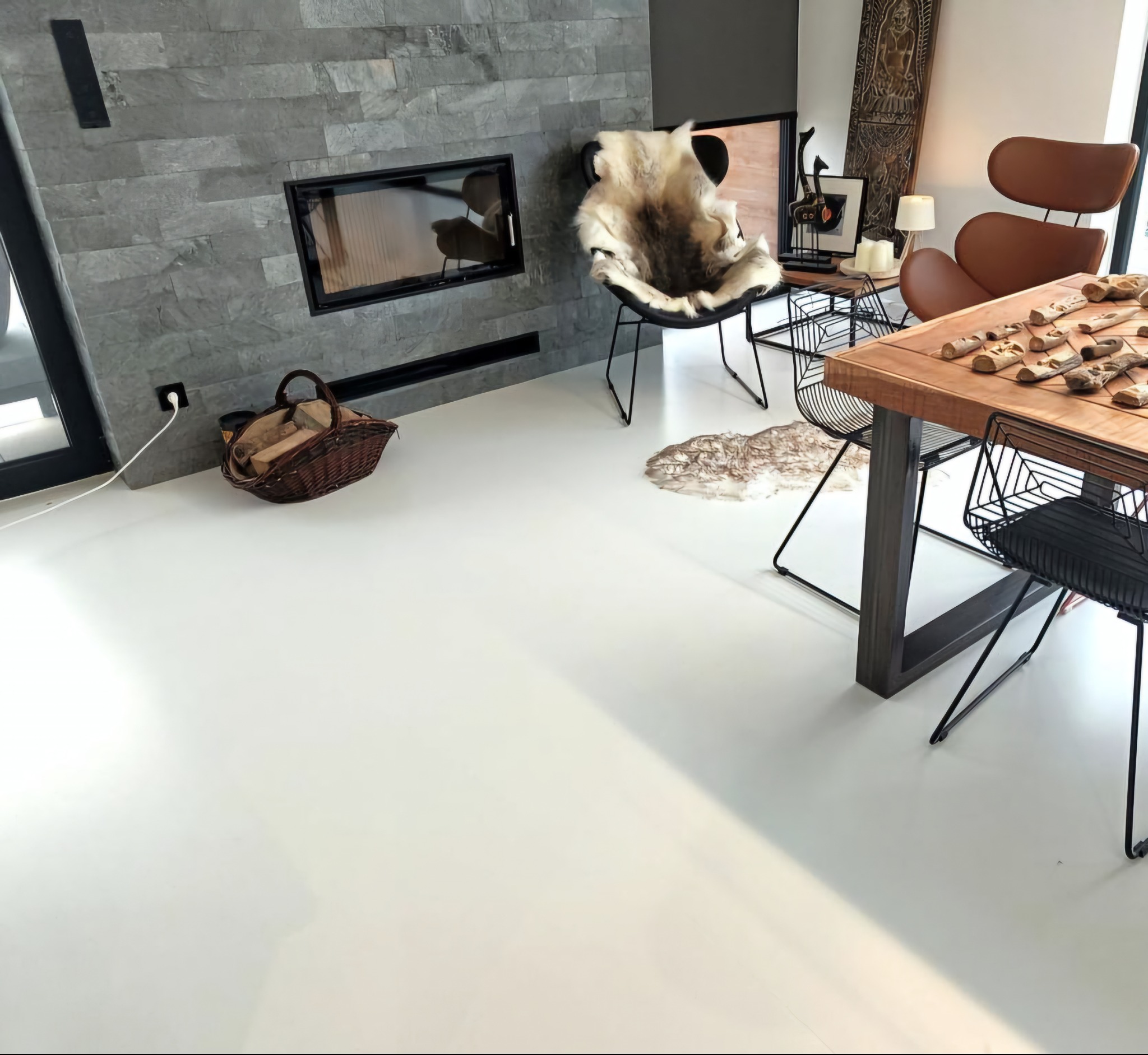 Biała, gładka posadzka w nowoczesnym salonie z kominkiem obłożonym szarym kamieniem. W tle designerskie krzesła i stół z drewnianym blatem.