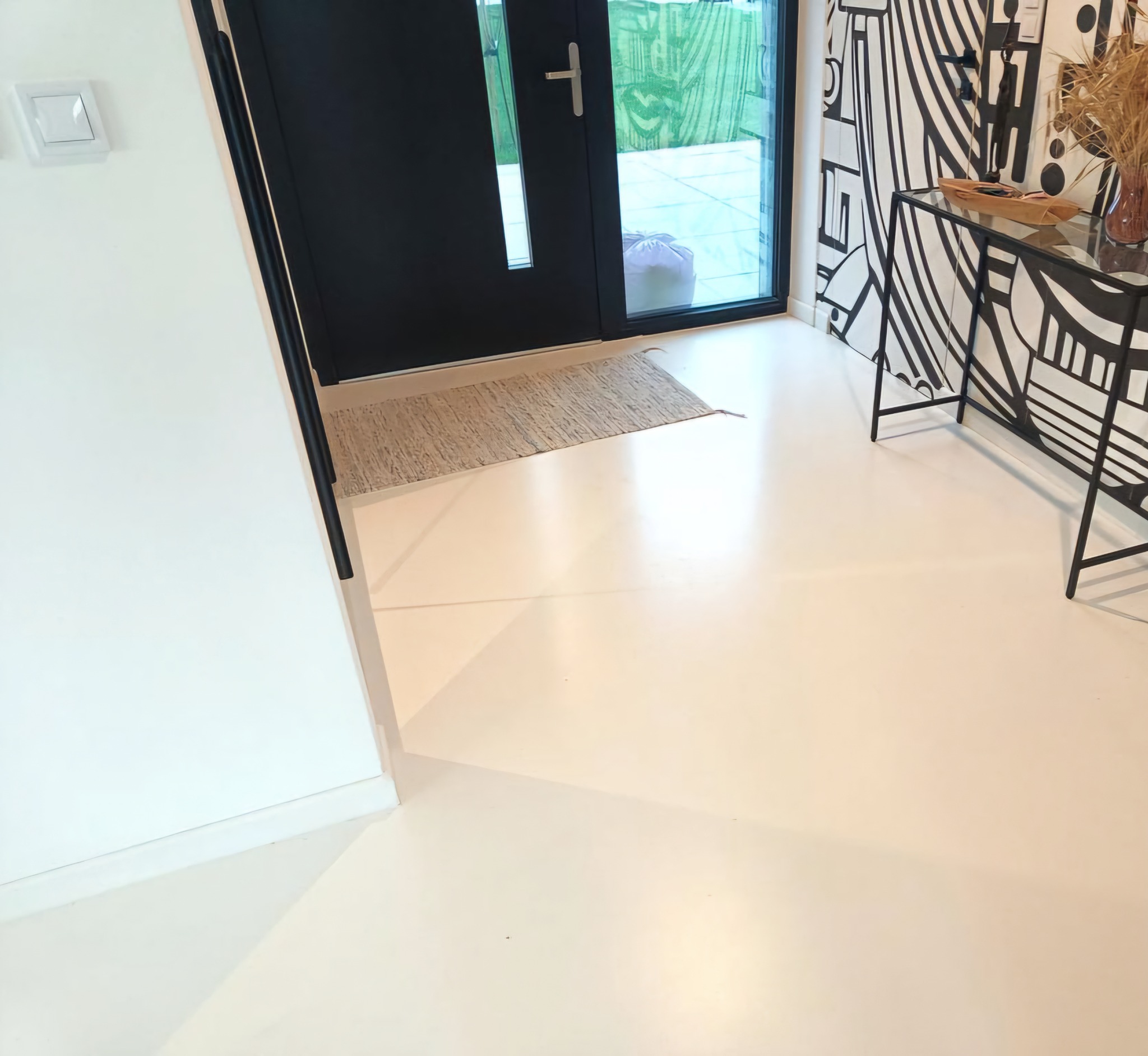 Jasna, jednolita posadzka żywiczna w nowoczesnym wnętrzu z czarnymi akcentami: drzwi, konsola i grafika na ścianie. Minimalistyczny dywanik przy wejściu.