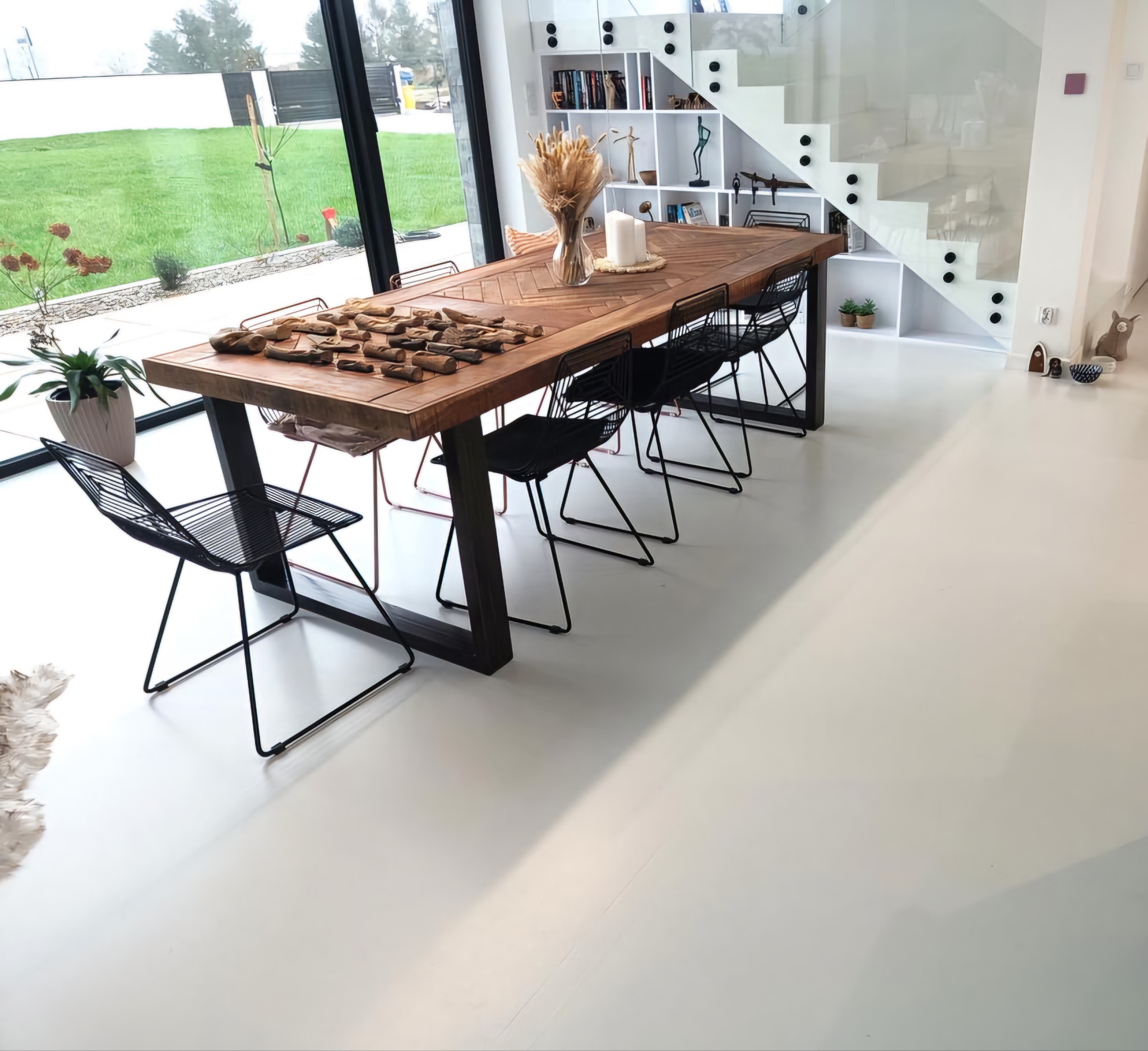 Jasne, białe wnętrze z podłogą żywiczną. Duży stół z drewnianym blatem i czarnymi krzesłami. Minimalistyczny design, duże okno z widokiem na zewnątrz.