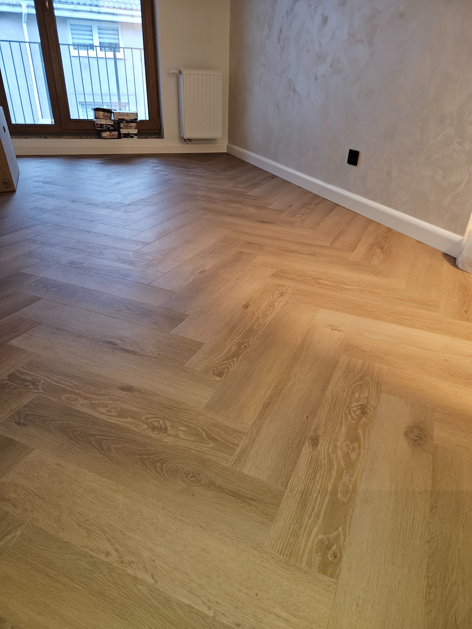 Jasne panele ułożone w jodełkę na podłodze w pokoju z oknem i grzejnikiem. Widoczne puszki po panelach przy oknie. Ściana wykończona w stylu dekoracyjnym.