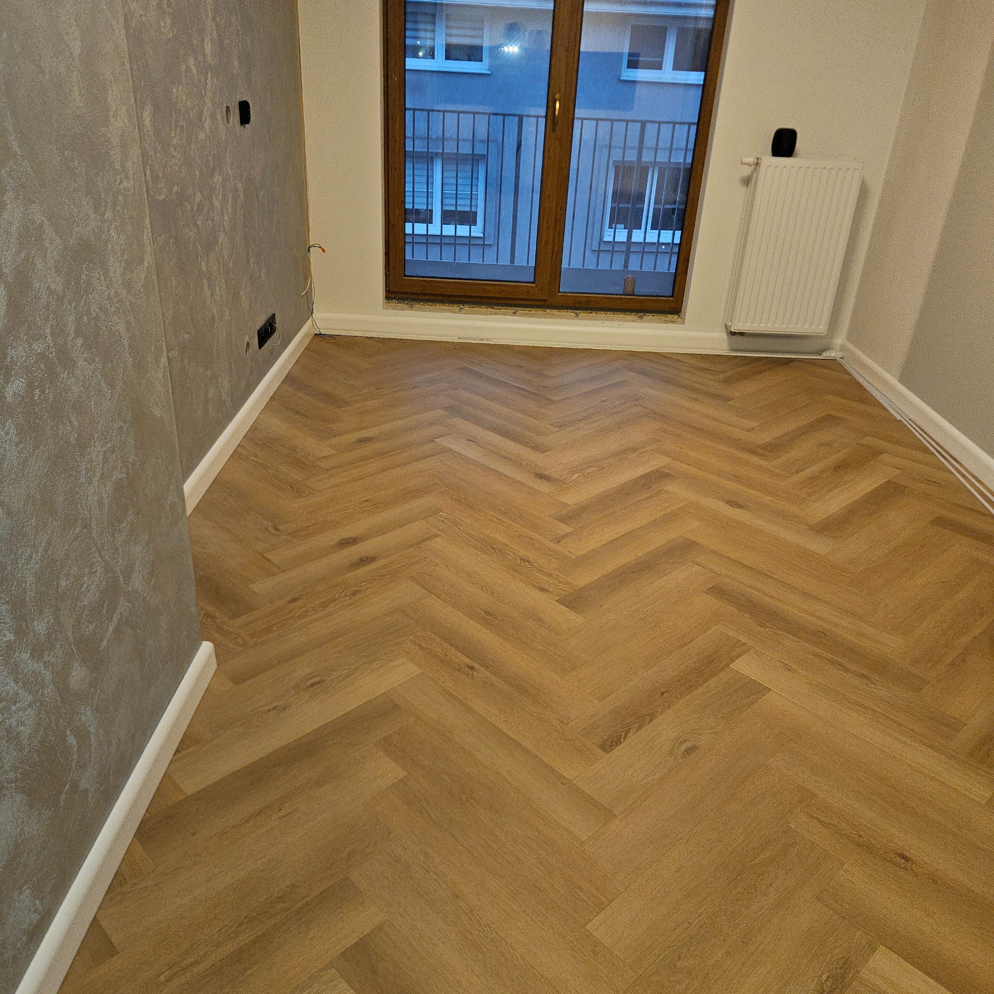 Jasne panele ułożone w jodełkę w pokoju z szarą ścianą i oknem balkonowym. Widoczny biały grzejnik i listwy przypodłogowe. Nowoczesne wykończenie wnętrza.