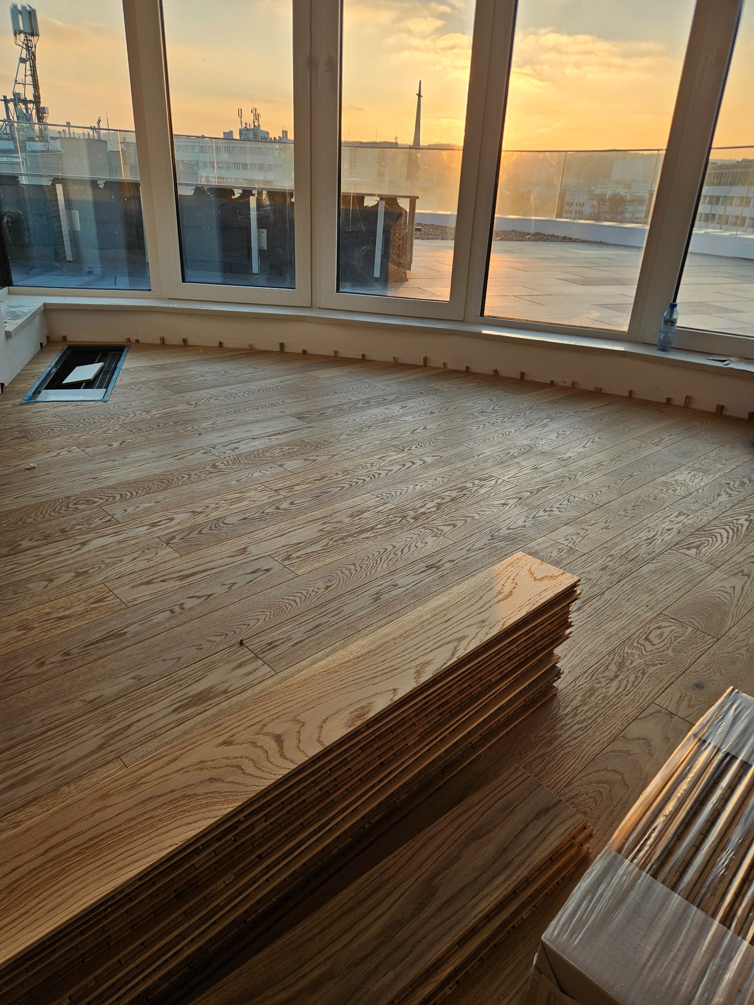 Układanie parkietu na balkonie z widokiem na miasto o zachodzie słońca. Część podłogi ułożona, widoczne paczki parkietu i listwy przypodłogowe. Otwór rewizyjny w podłodze.