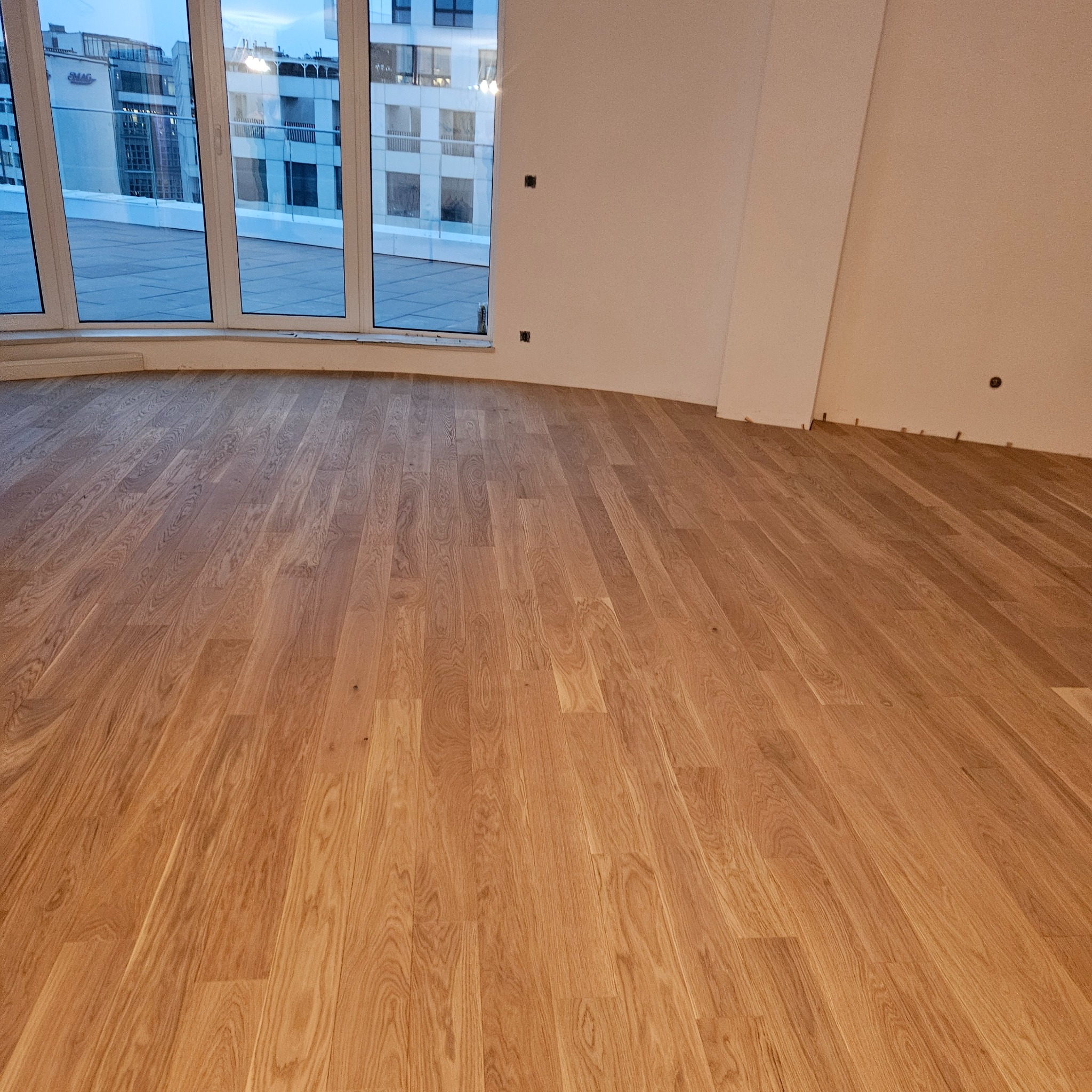 Jasny parkiet ułożony w nowym apartamencie z dużymi oknami balkonowymi. Widoczne listwy przypodłogowe i fragmenty ścian przygotowane do malowania.