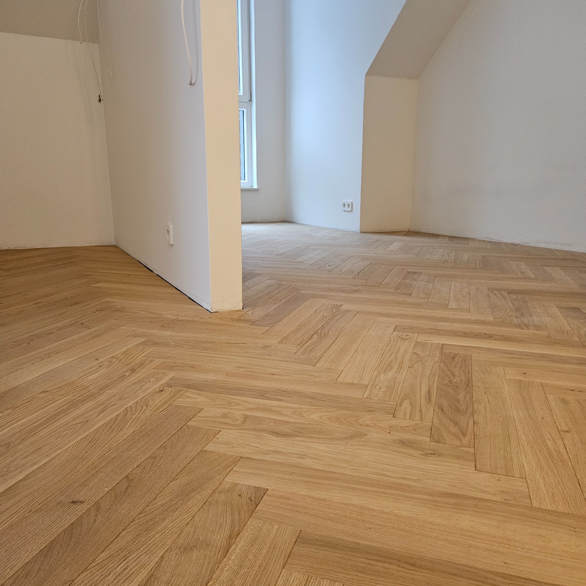 Jasny parkiet ułożony we wzór jodełki francuskiej w przestronnym, jasnym wnętrzu. Widoczne białe ściany i fragment okna. Naturalne oświetlenie podkreśla strukturę drewna.