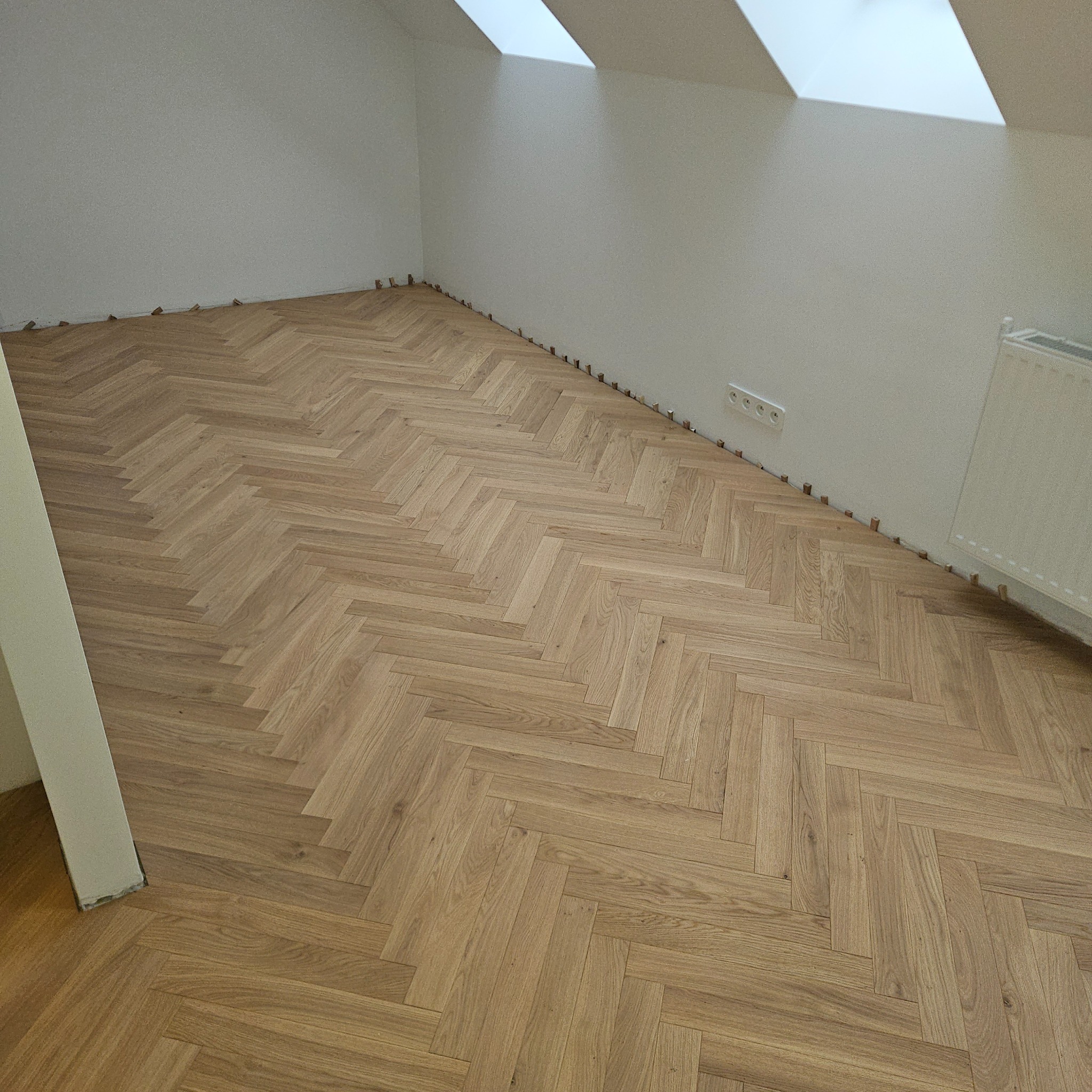Ułożony parkiet w jodełkę w jasnym odcieniu drewna, widoczne kliny dystansowe przy ścianie, pokój na poddaszu z oknami dachowymi.