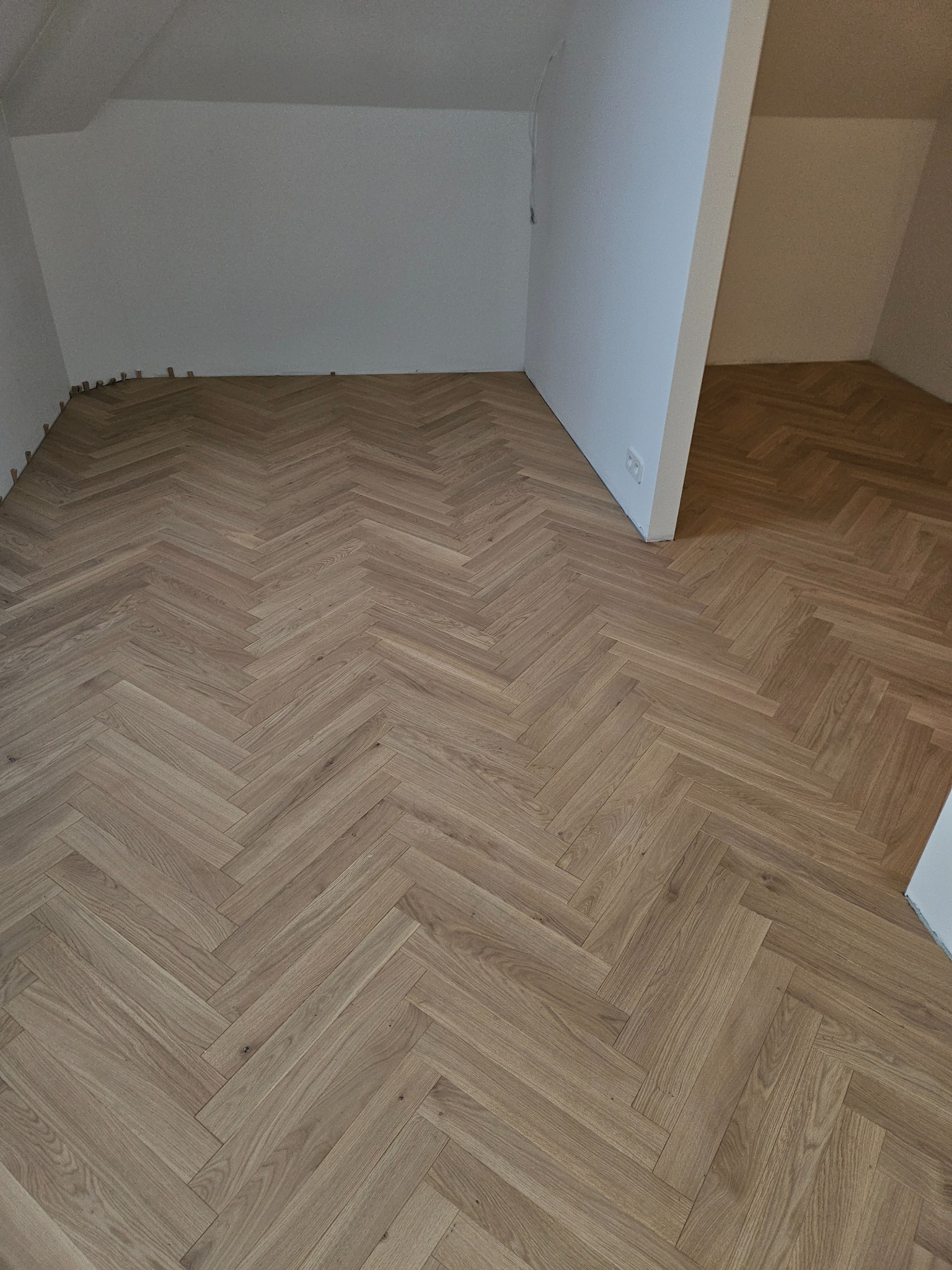 Jasny parkiet ułożony we wzór jodełki francuskiej w pokoju na poddaszu. Widoczne listwy przypodłogowe i fragment drugiego pomieszczenia.