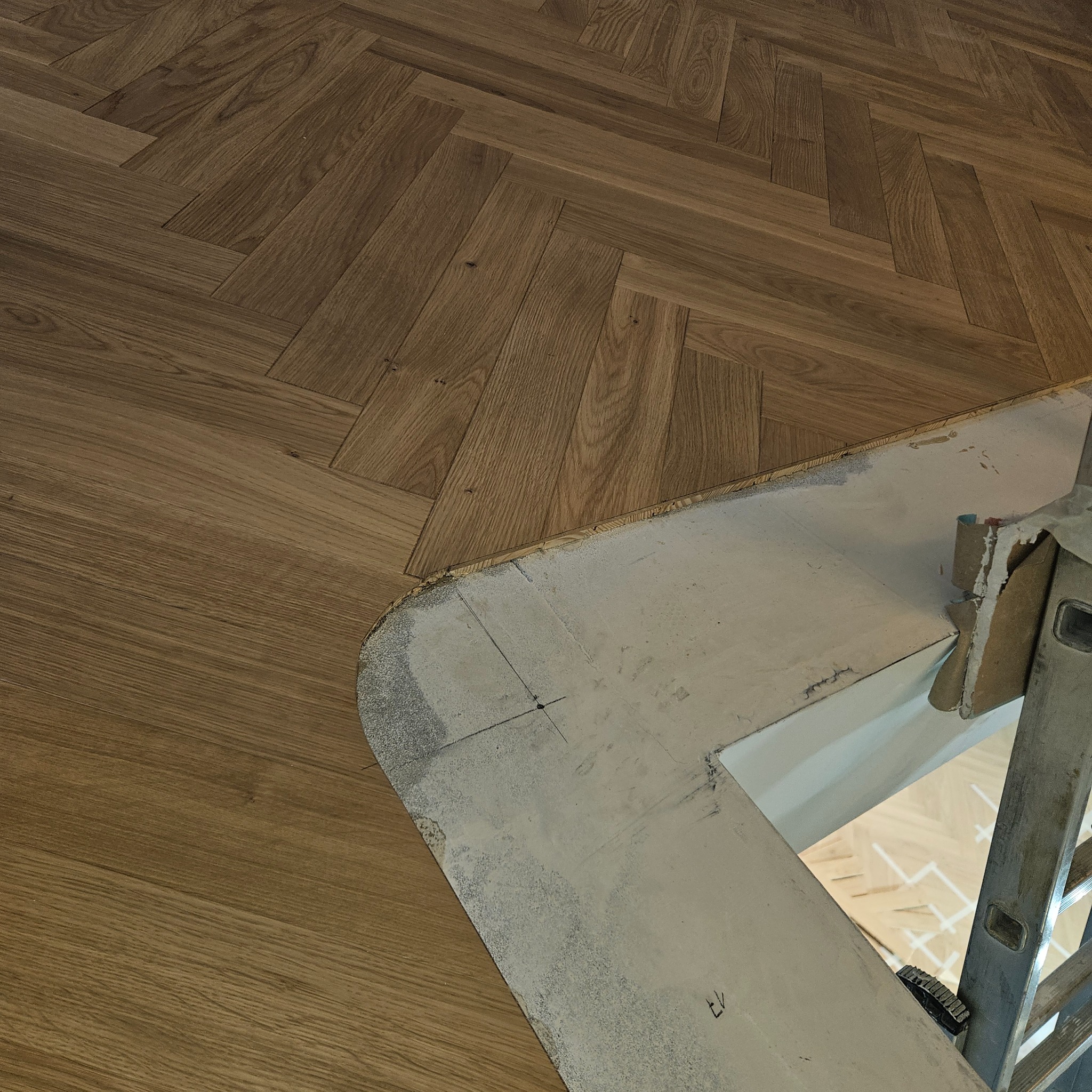 Układanie parkietu w jodełkę w Kartuzach. Widoczny fragment podłogi i szablon do wykończenia krawędzi. Na podłodze stoi drabina.