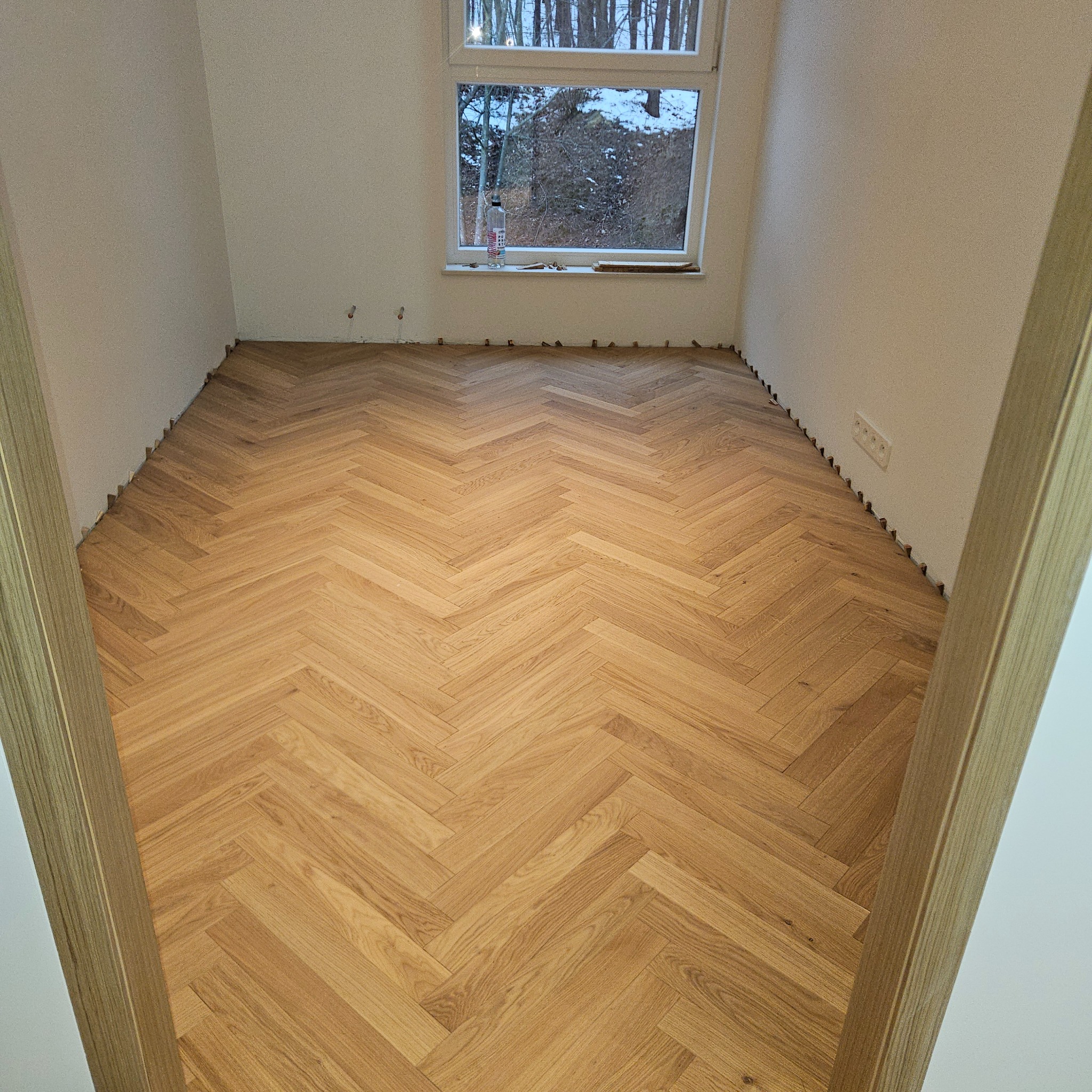 Ułożony parkiet w jodełkę w jasnym pomieszczeniu z oknem. Widoczne listwy dystansowe przy ścianach, sugerujące etap prac wykończeniowych. Naturalne światło dzienne.