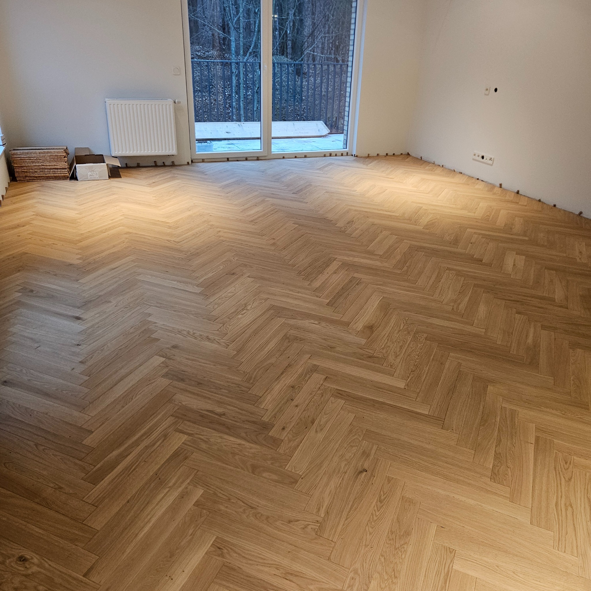 Ułożony parkiet w jodełkę w jasnym odcieniu drewna, widoczne listwy przypodłogowe i okno balkonowe z widokiem na drzewa. Częściowo ułożone deski parkietu w rogu pomieszczenia.