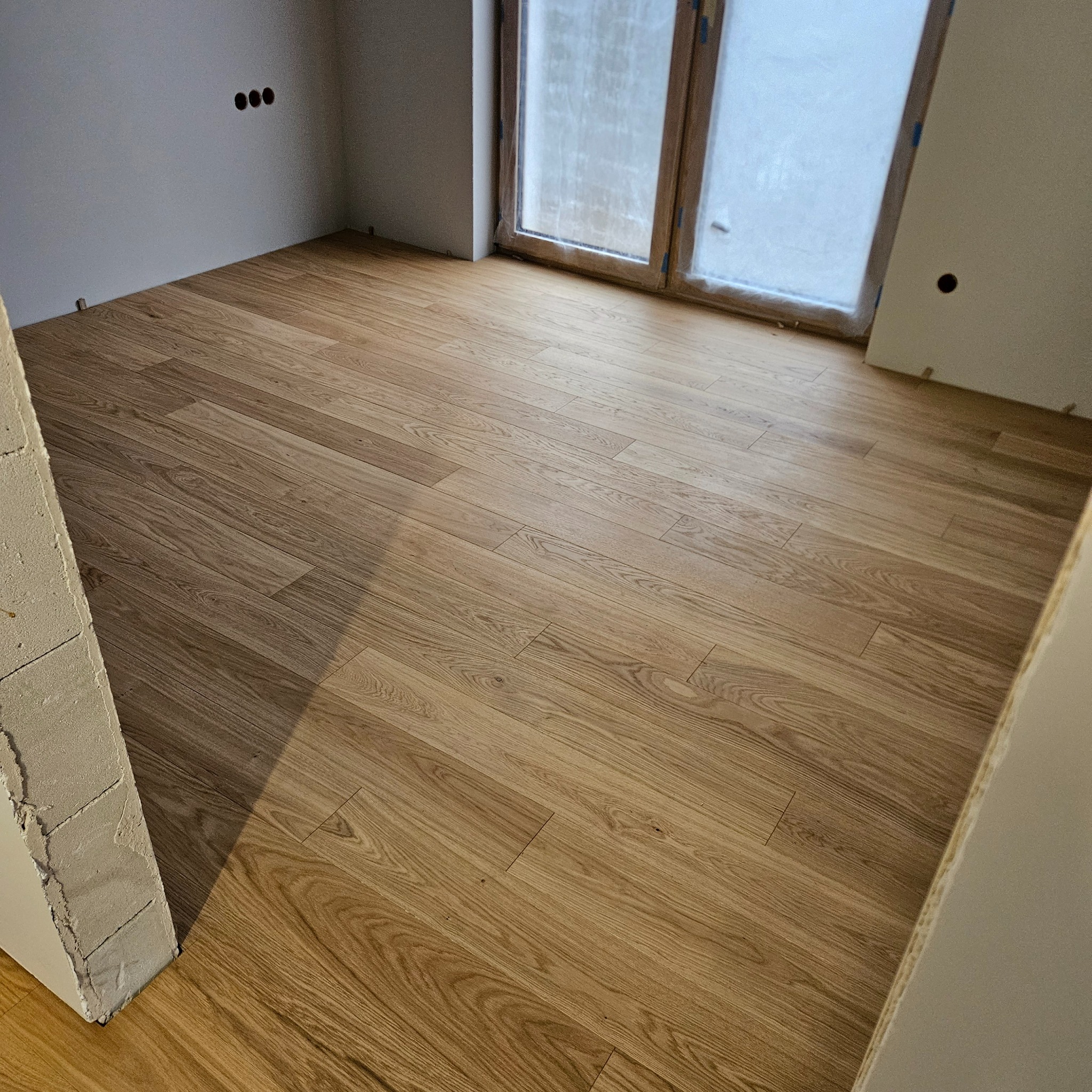 Jasne panele podłogowe ułożone w pokoju z oknem. Widoczne ślady po remoncie na ścianach i framugach. Naturalne światło podkreśla strukturę drewna.