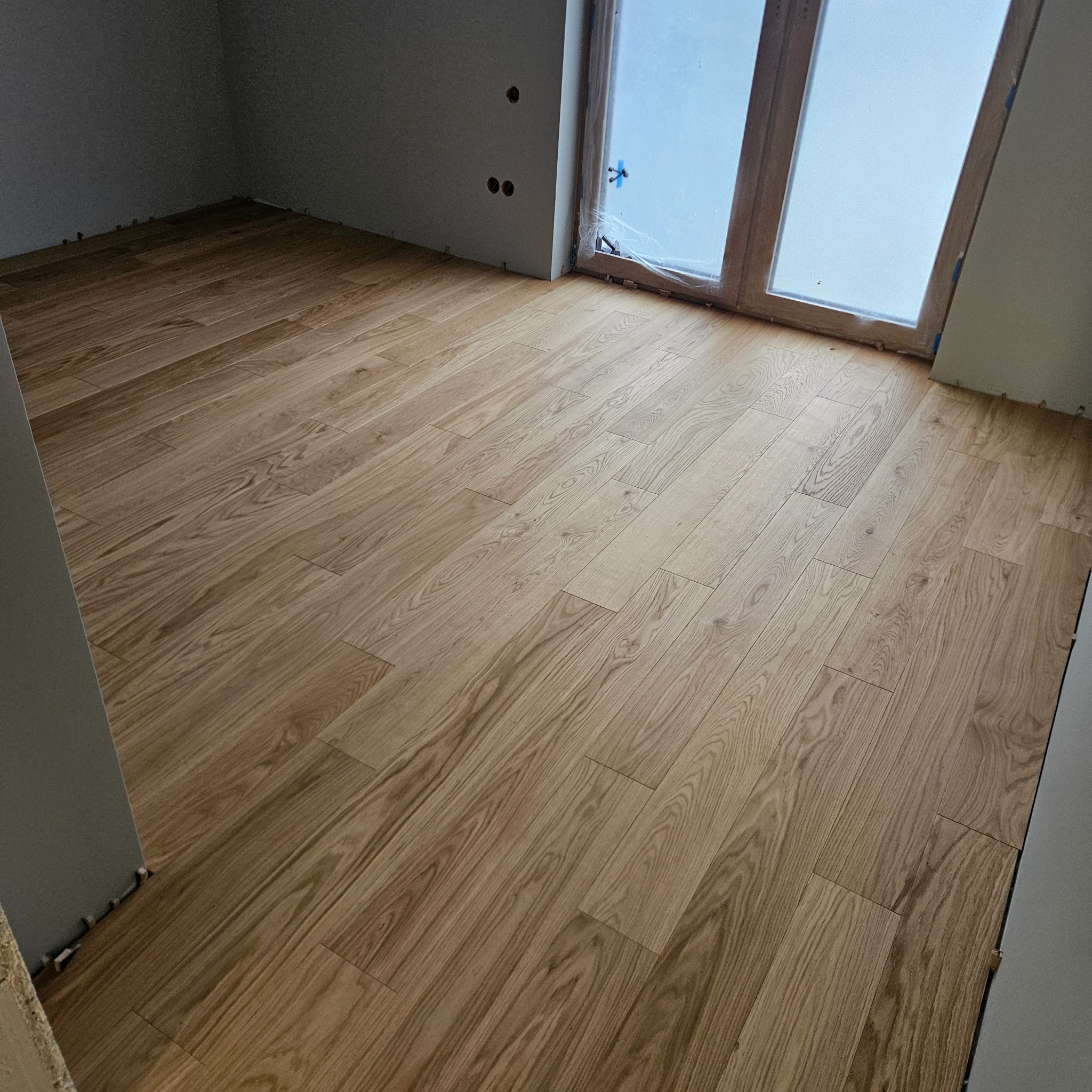 Ułożony parkiet w jasnym odcieniu drewna w nowym pomieszczeniu z oknem. Widoczne listwy przypodłogowe i przygotowane otwory na gniazdka elektryczne.