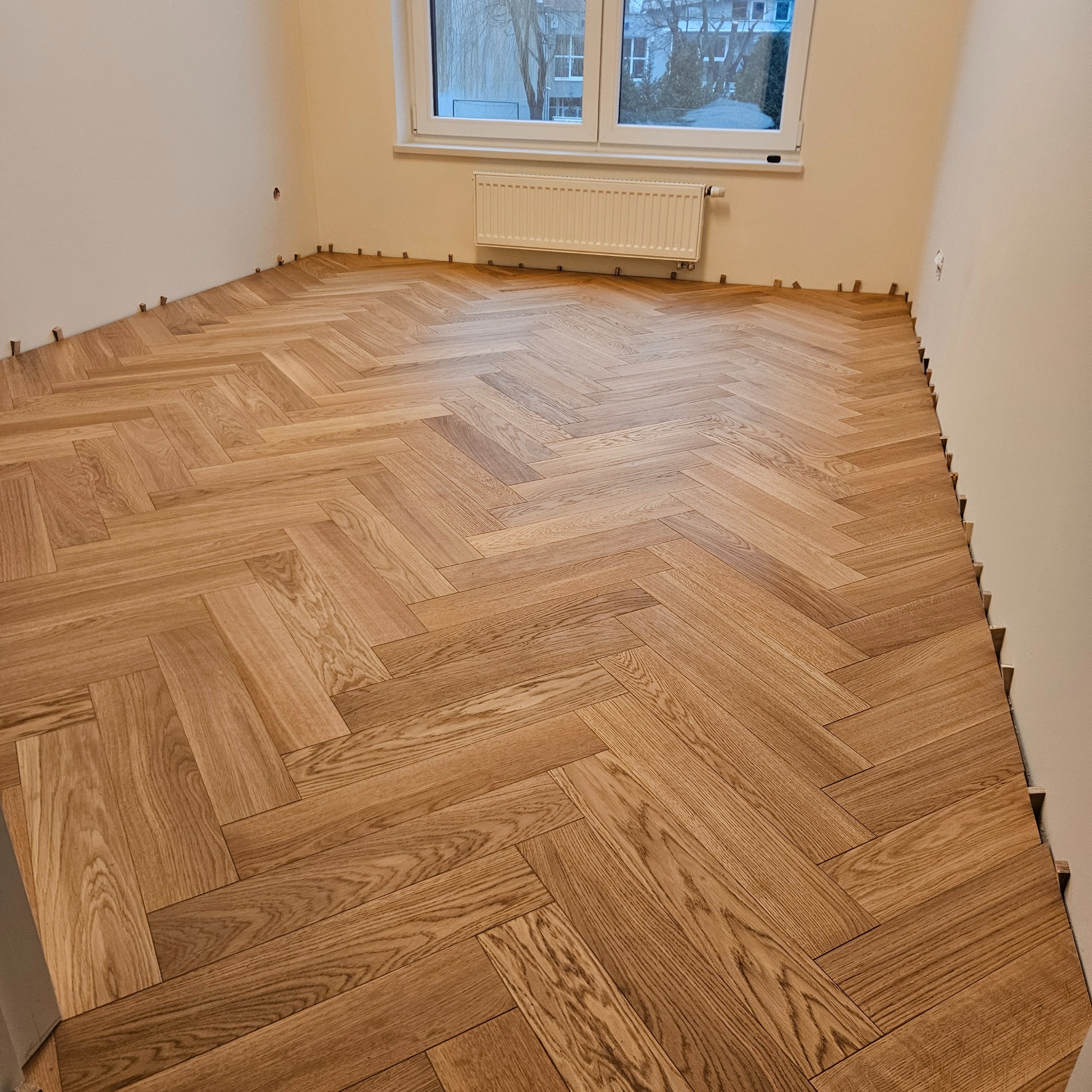 Układanie parkietu w jodełkę w jasnym pokoju z oknem. Widoczne kliny dystansowe przy ścianach. Parkiet o naturalnym odcieniu drewna, ułożony w klasyczny wzór.