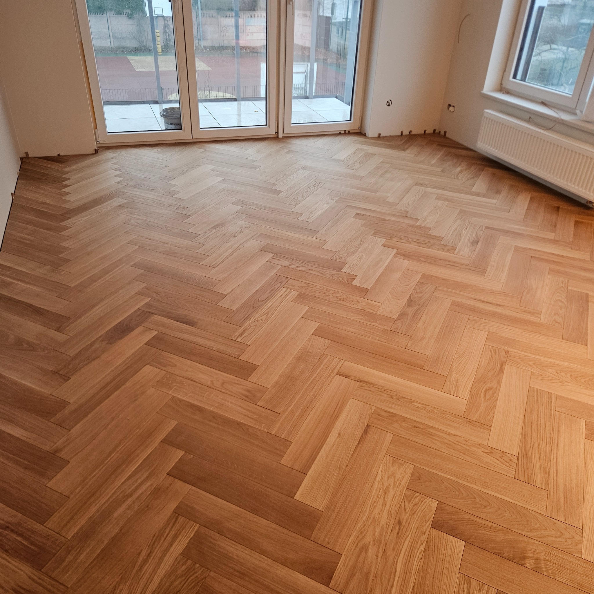 Ułożony parkiet w jodełkę francuską w jasnym pomieszczeniu z dużymi oknami balkonowymi. Widoczne listwy przypodłogowe i grzejnik. Naturalne oświetlenie.