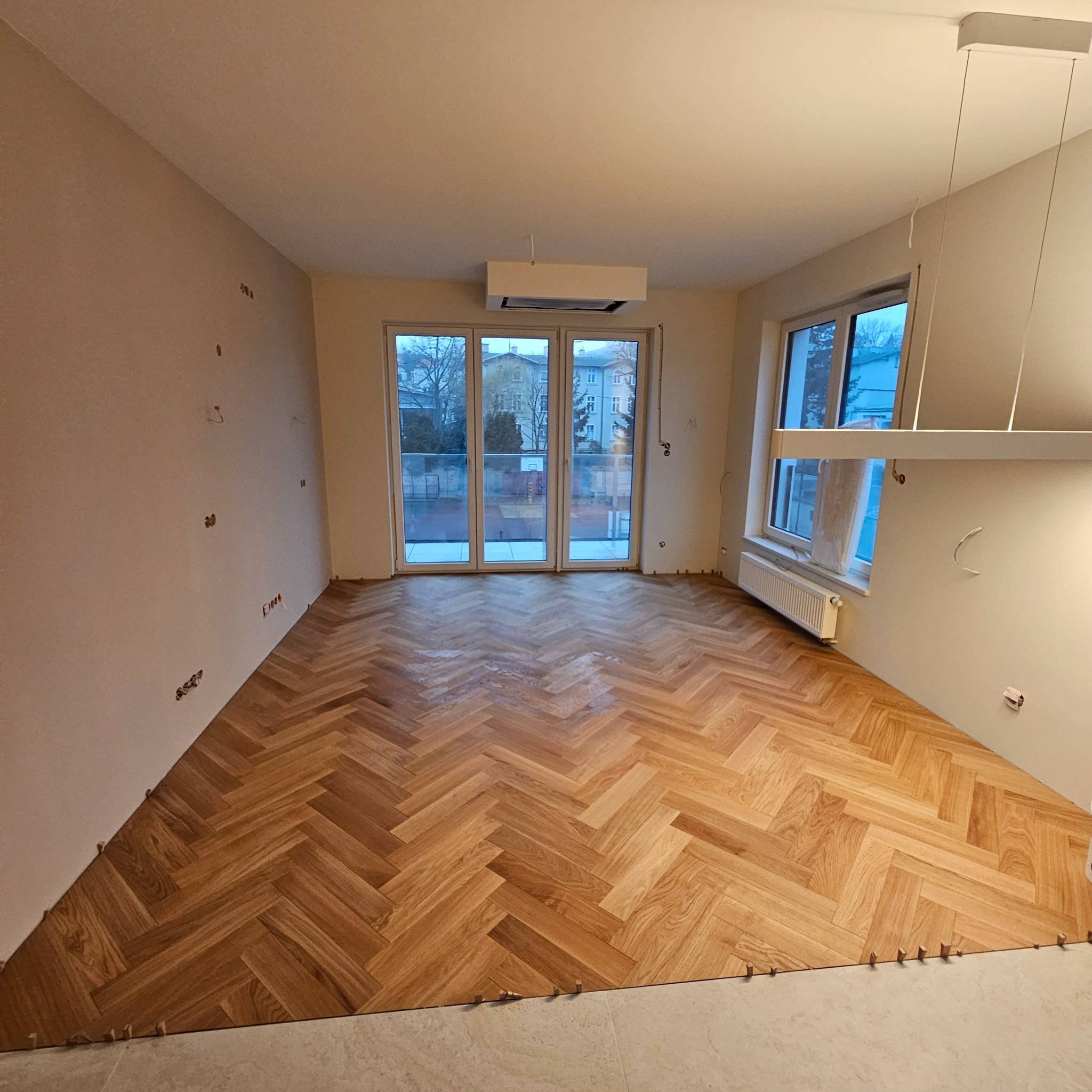 Jasny parkiet ułożony we wzór jodełki w przestronnym pokoju z dużymi oknami balkonowymi. Widoczne listwy dystansowe przy ścianie, surowy stan ścian przed wykończeniem.