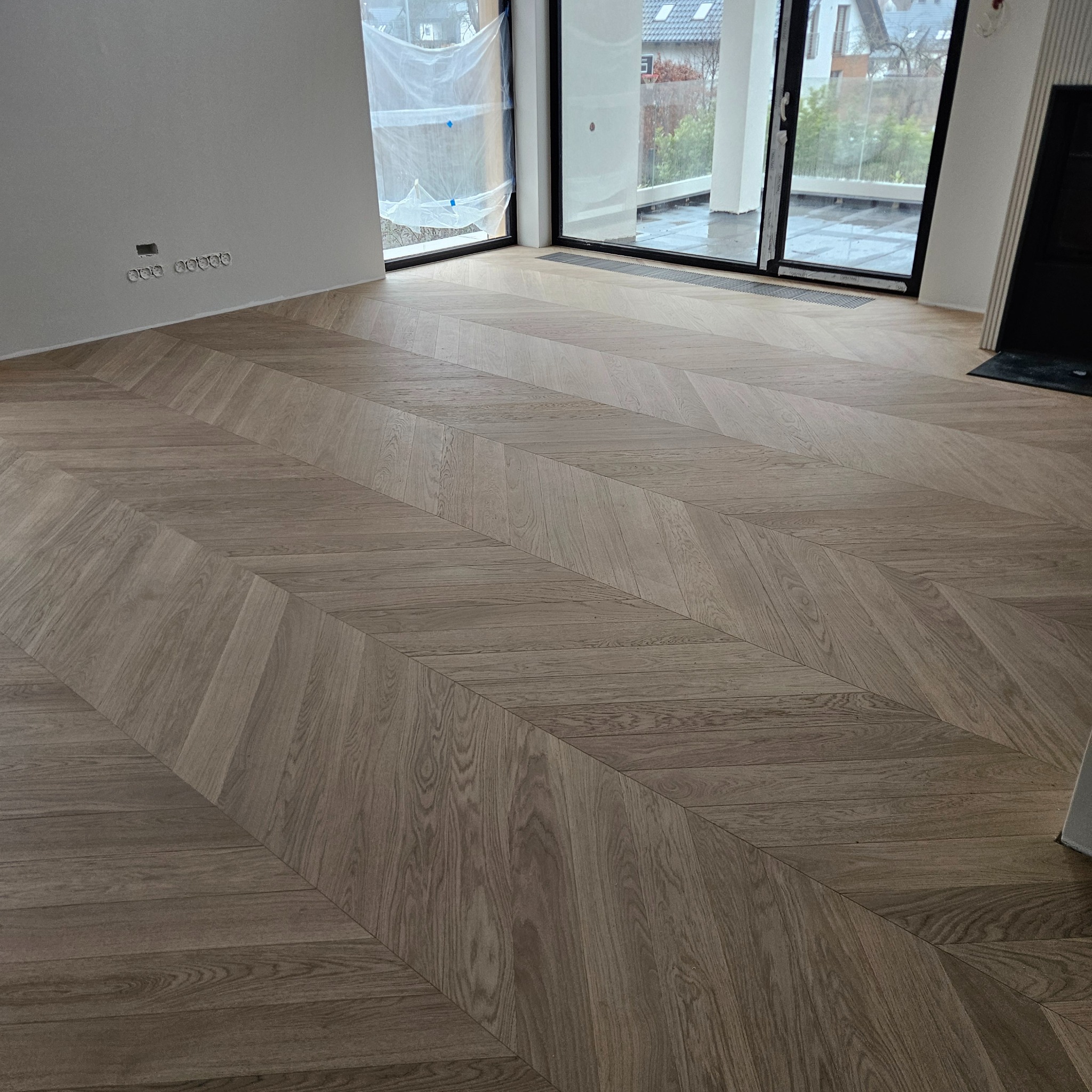 Ukończona podłoga z parkietu ułożonego we wzór jodełki w jasnym odcieniu drewna. Widoczne duże okna z widokiem na zewnątrz i fragment ściany z gniazdkami elektrycznymi.