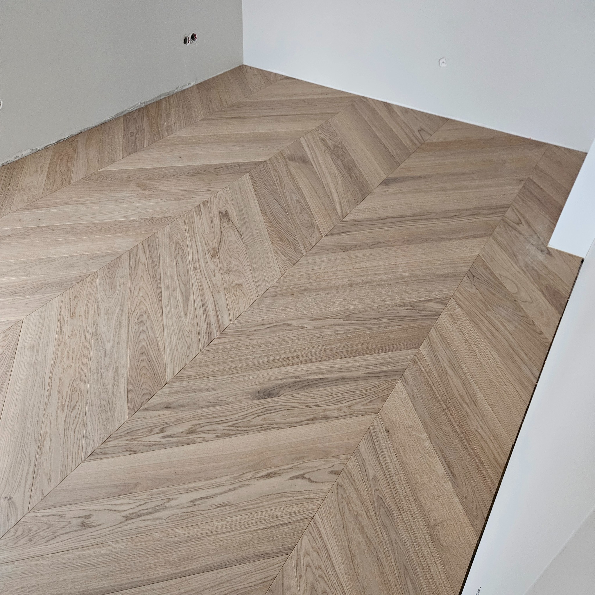 Ukończona podłoga z jasnego drewna ułożona we wzór jodełki francuskiej w nowym wnętrzu. Widoczne gniazdka elektryczne w ścianie.