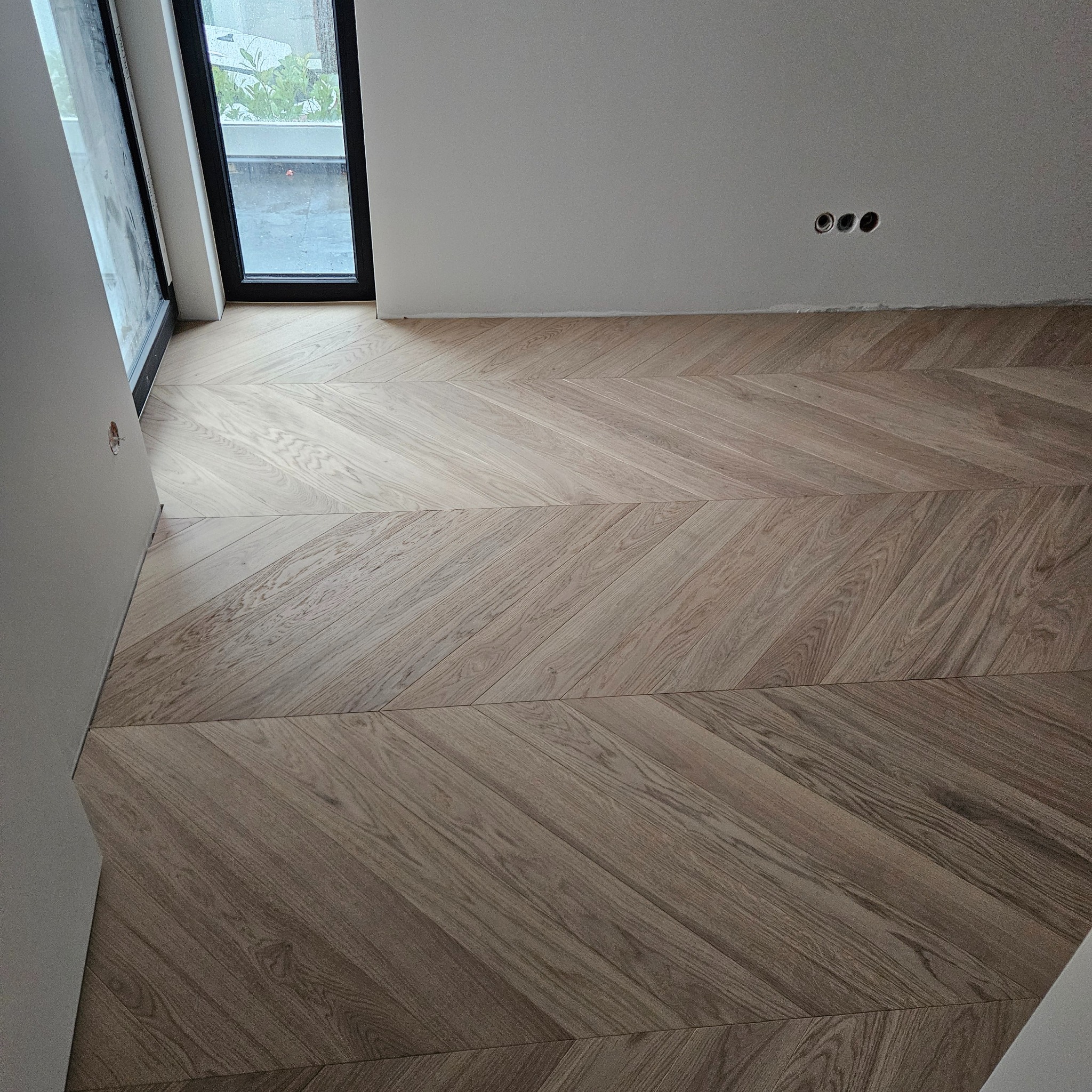 Ułożony parkiet w jodełkę francuską, jasne drewno, widok na fragment okna i białą ścianę z trzema gniazdkami. Nowoczesne wykończenie podłogi.